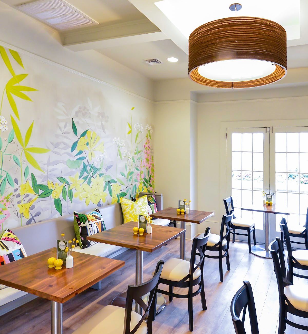 Bright café interior with wooden tables, black chairs, floral wall mural, and pendant light