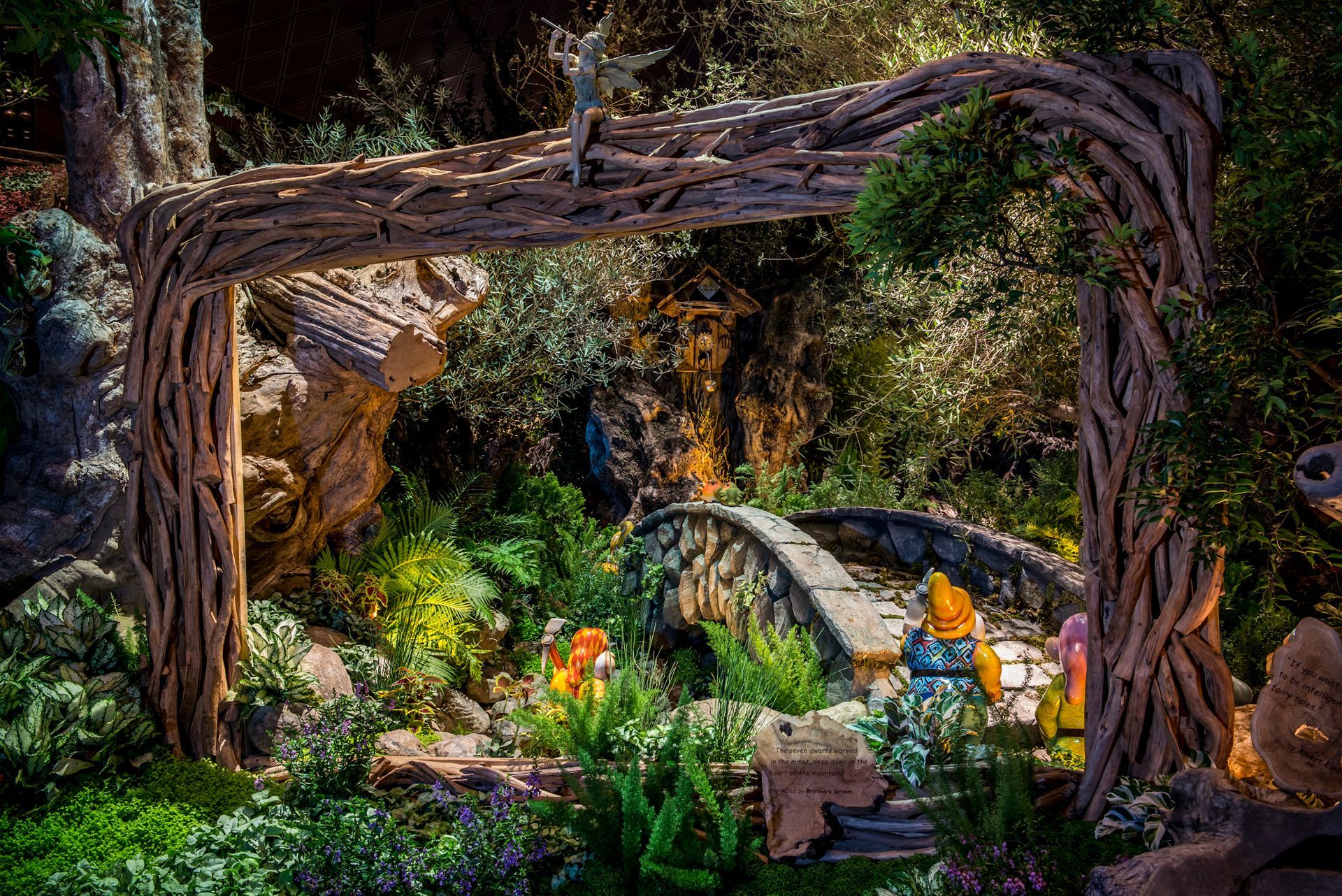 A whimsical garden scene with a stone bridge, archway, and small figures amidst lush greenery.