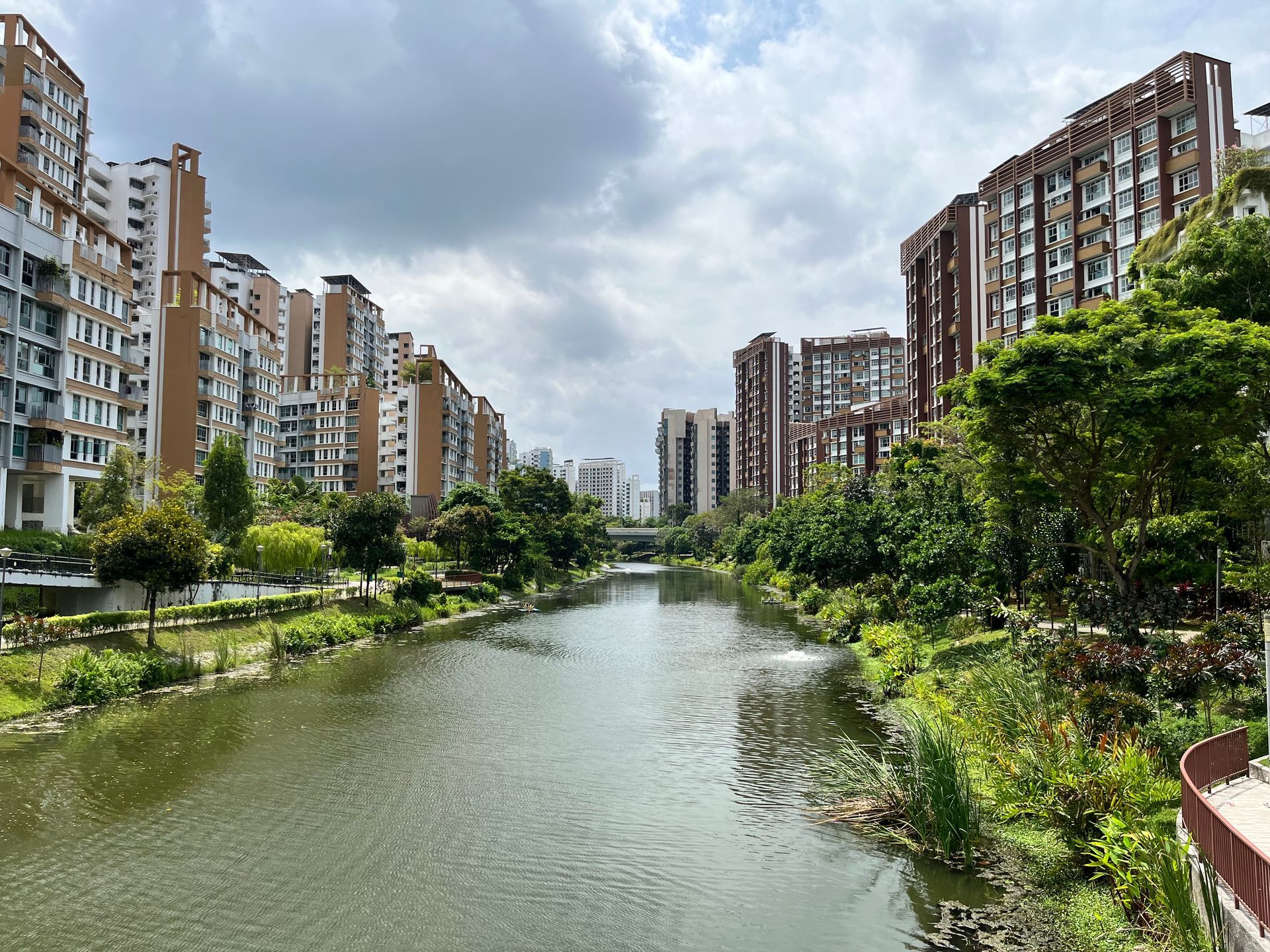 A river flows through a residential area lined with tall buildings and lush greenery under a cloudy sky.