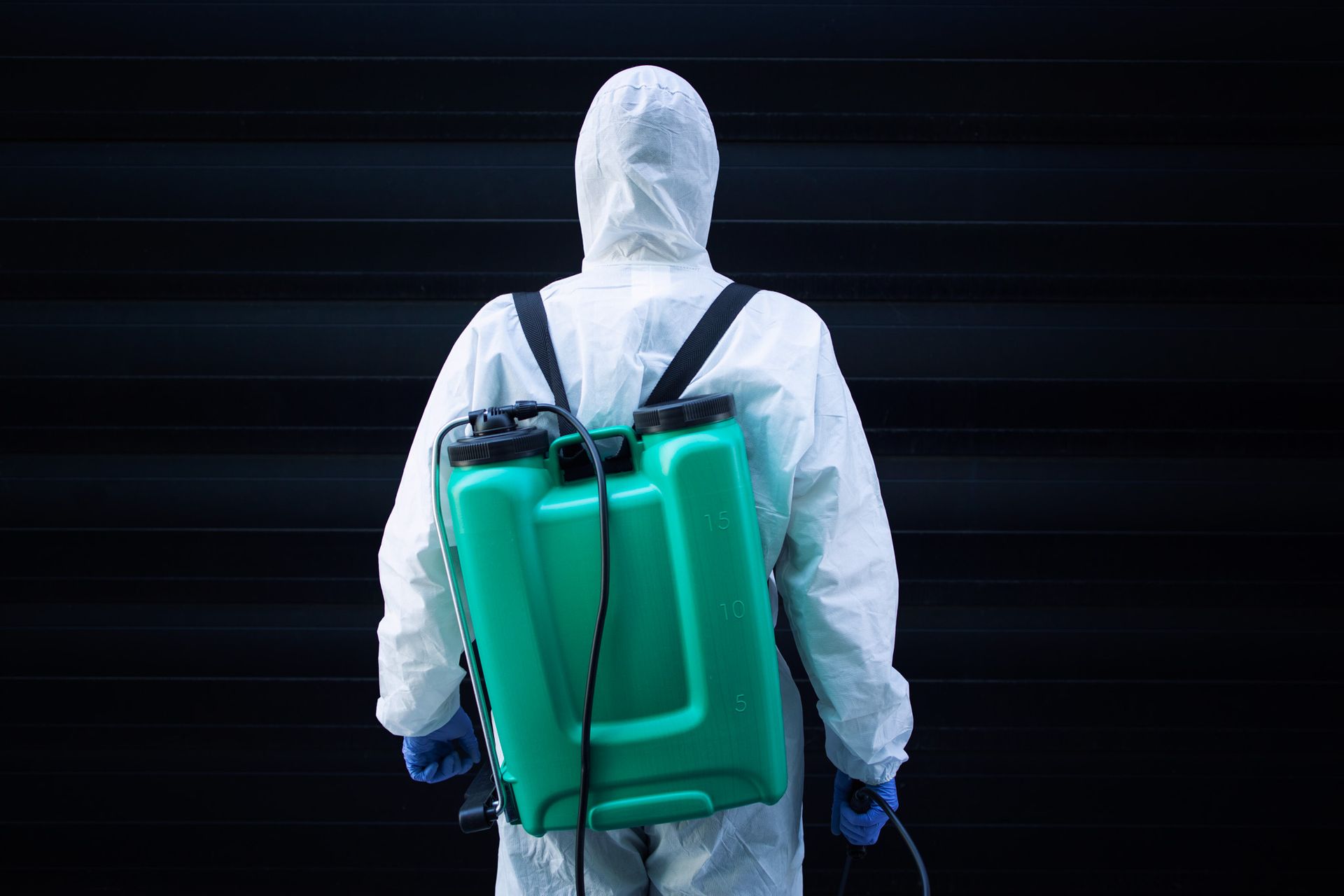 Person in white hazmat suit wearing a teal backpack sprayer against a dark background