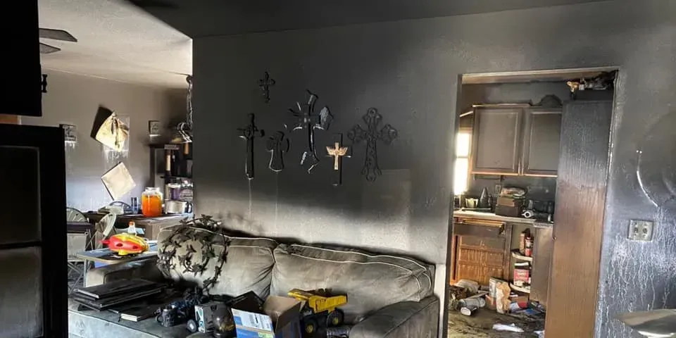 Cluttered living room with a worn sofa, wall decor, and a doorway to a messy kitchen area