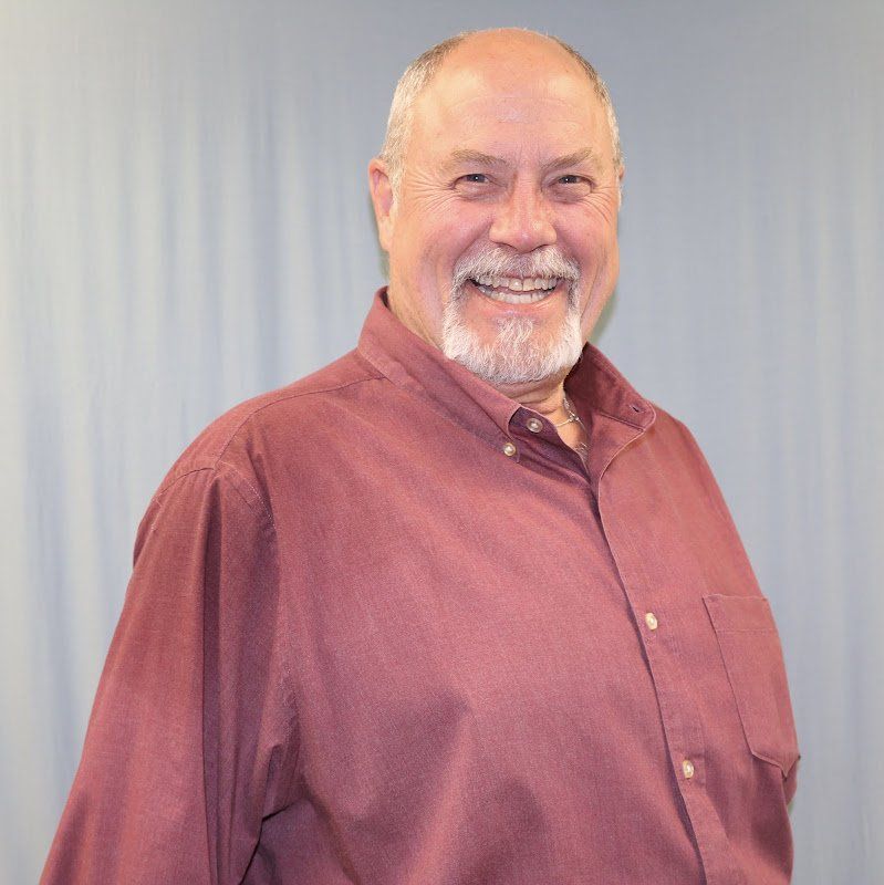 Man with a white beard smiling, wearing a maroon button-down shirt. Blue background.