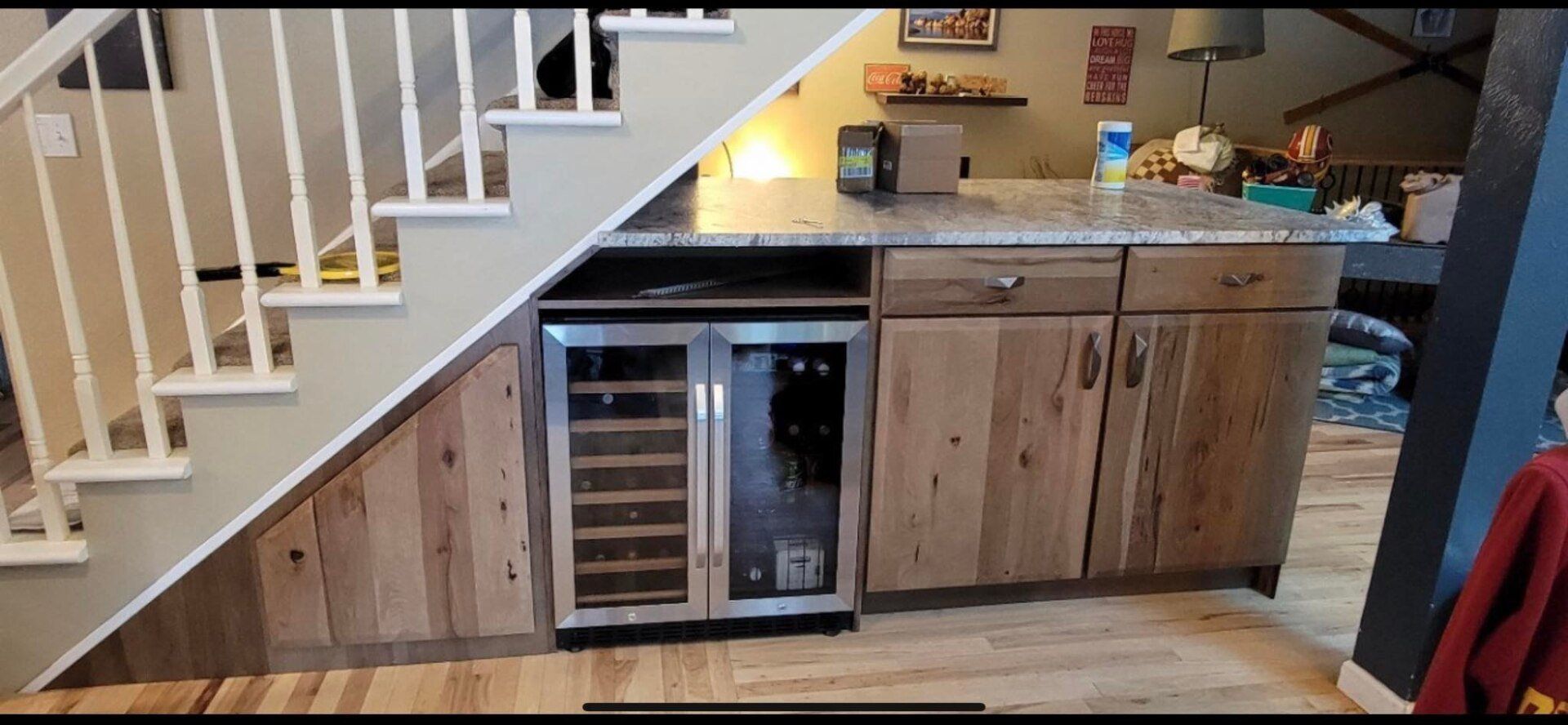 Kitchen island with a wine fridge, beneath a staircase. Wooden cabinets and hardwood flooring.