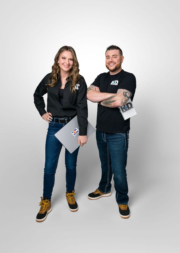 Woman and man standing, smiling. Woman holds a laptop, man holds papers, both wearing black shirts and jeans.