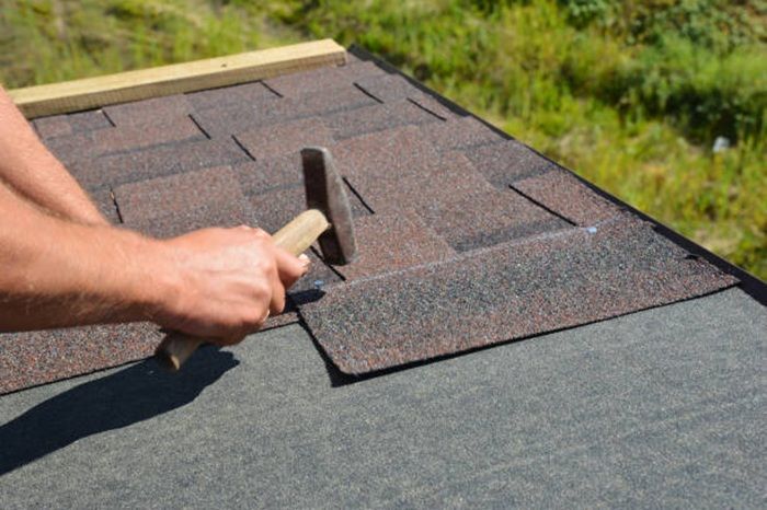 Person hammering asphalt shingles on a roof.