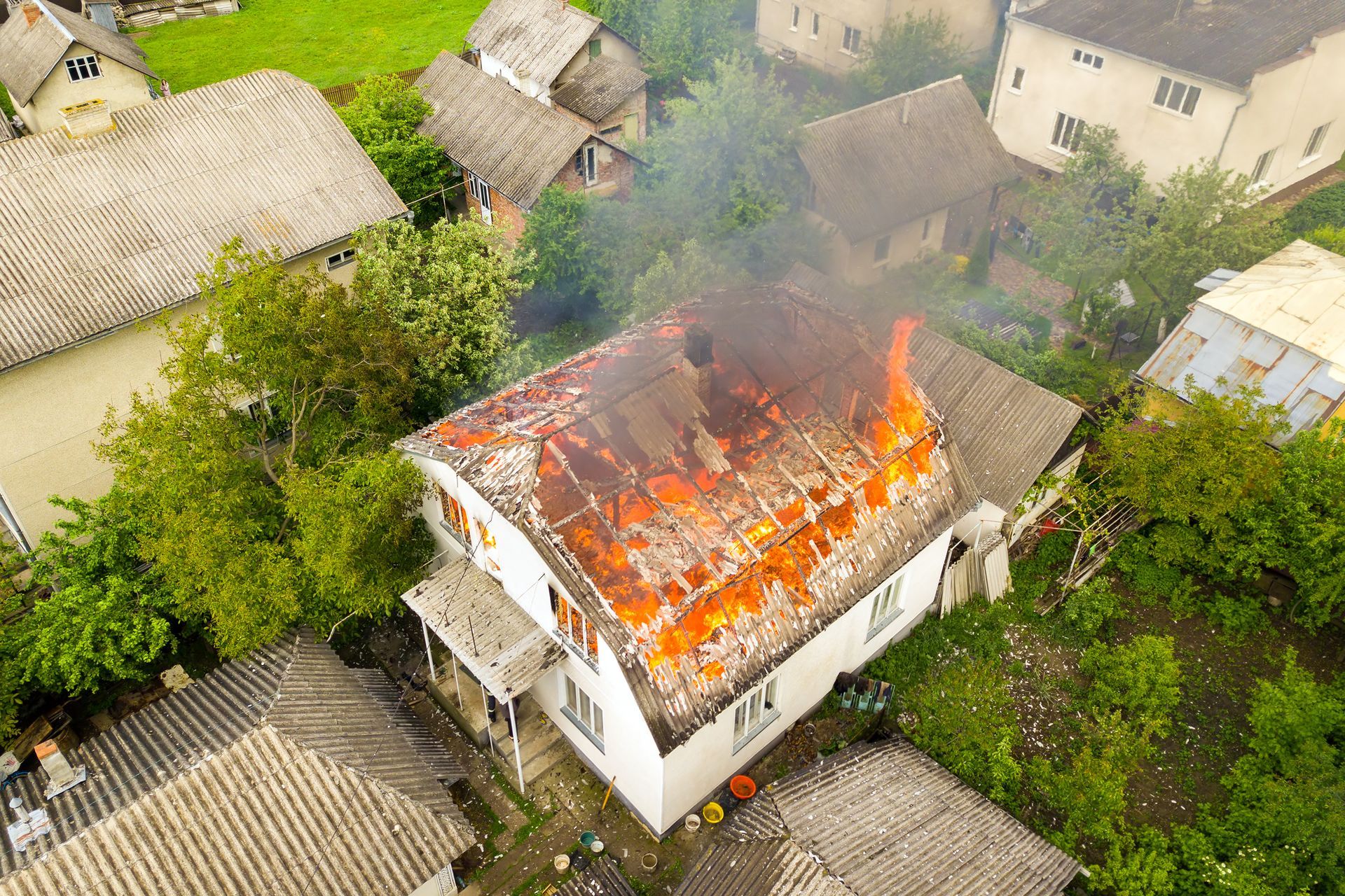 Overhead view of a house on fire, flames erupting from the roof. Surrounding buildings and trees are visible.