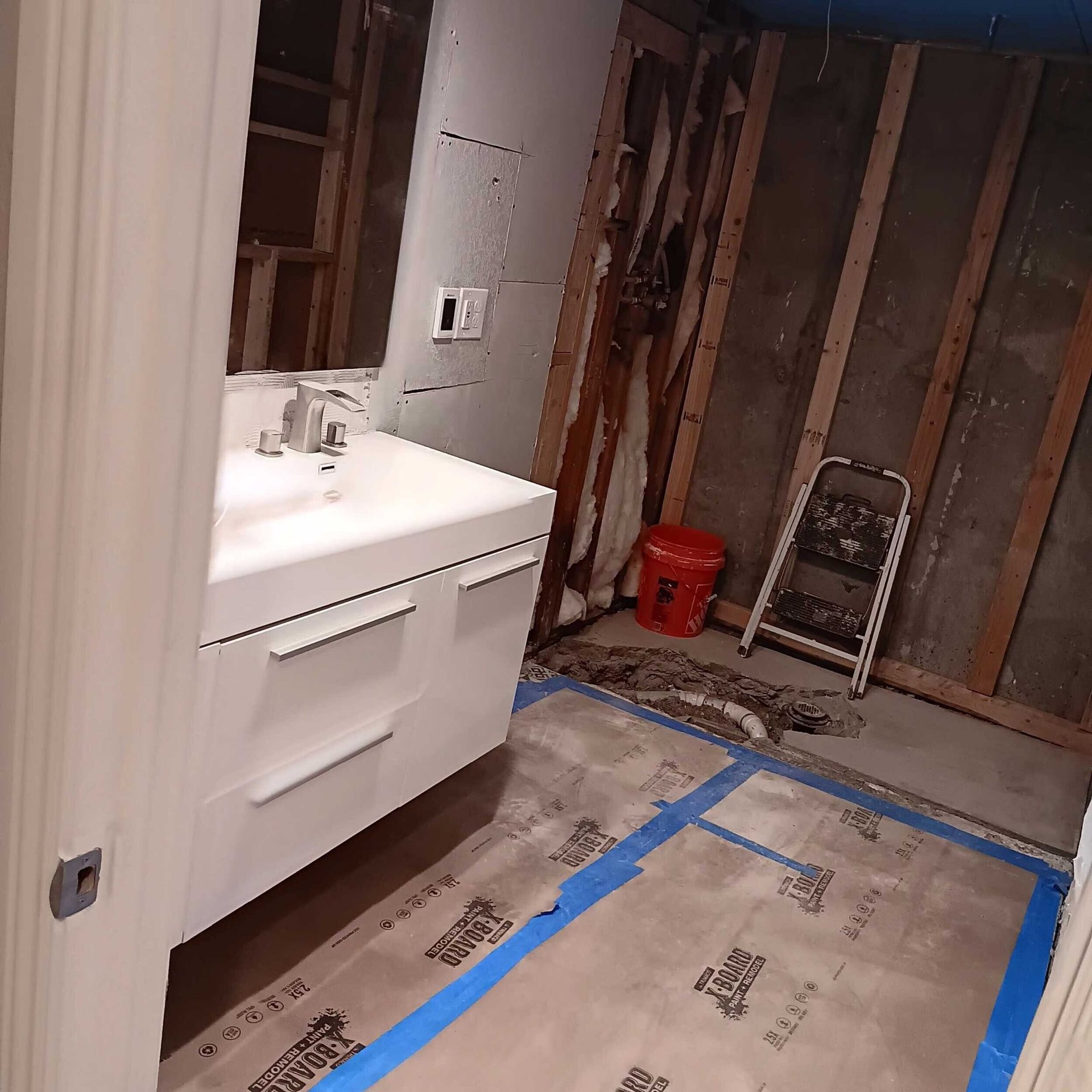 Bathroom under construction: White vanity, exposed wall studs, red bucket, ladder, and floor protection in place.
