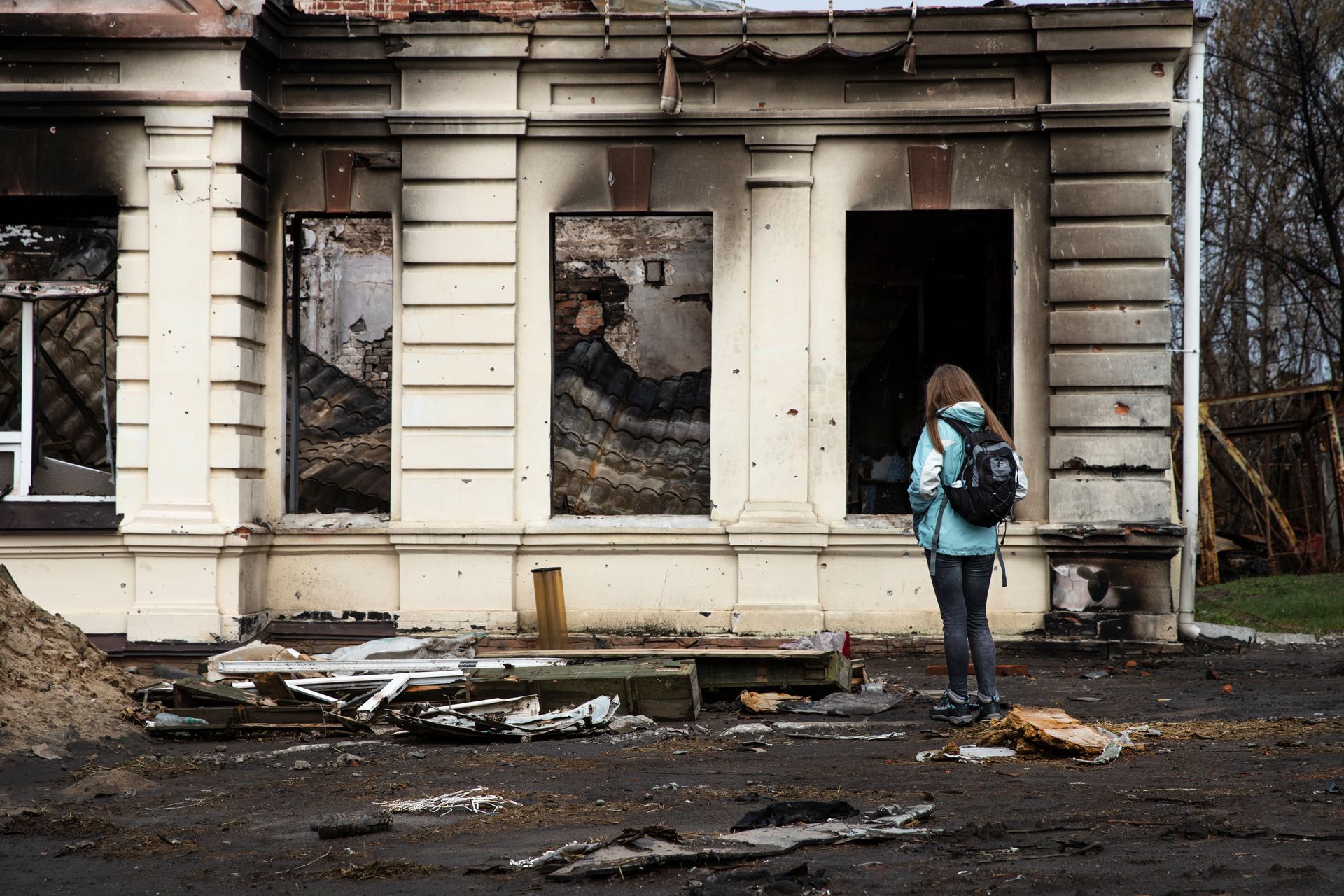 Woman looking at a damaged building facade with burned-out windows; debris on the ground.