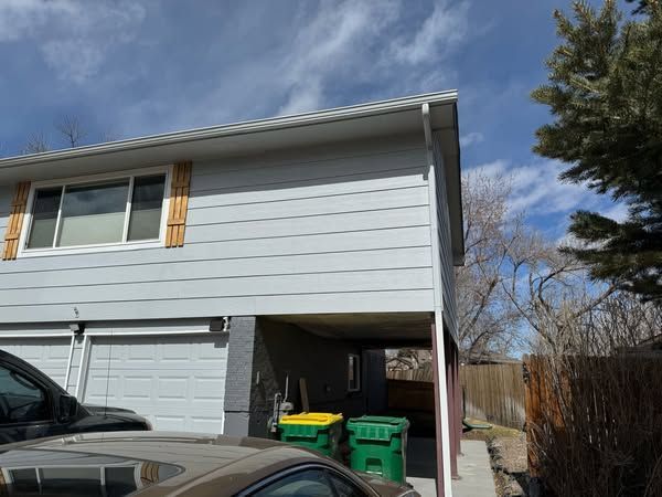 Two-story house with light blue siding, white trim, and a garage. Two green bins are beneath a carport.