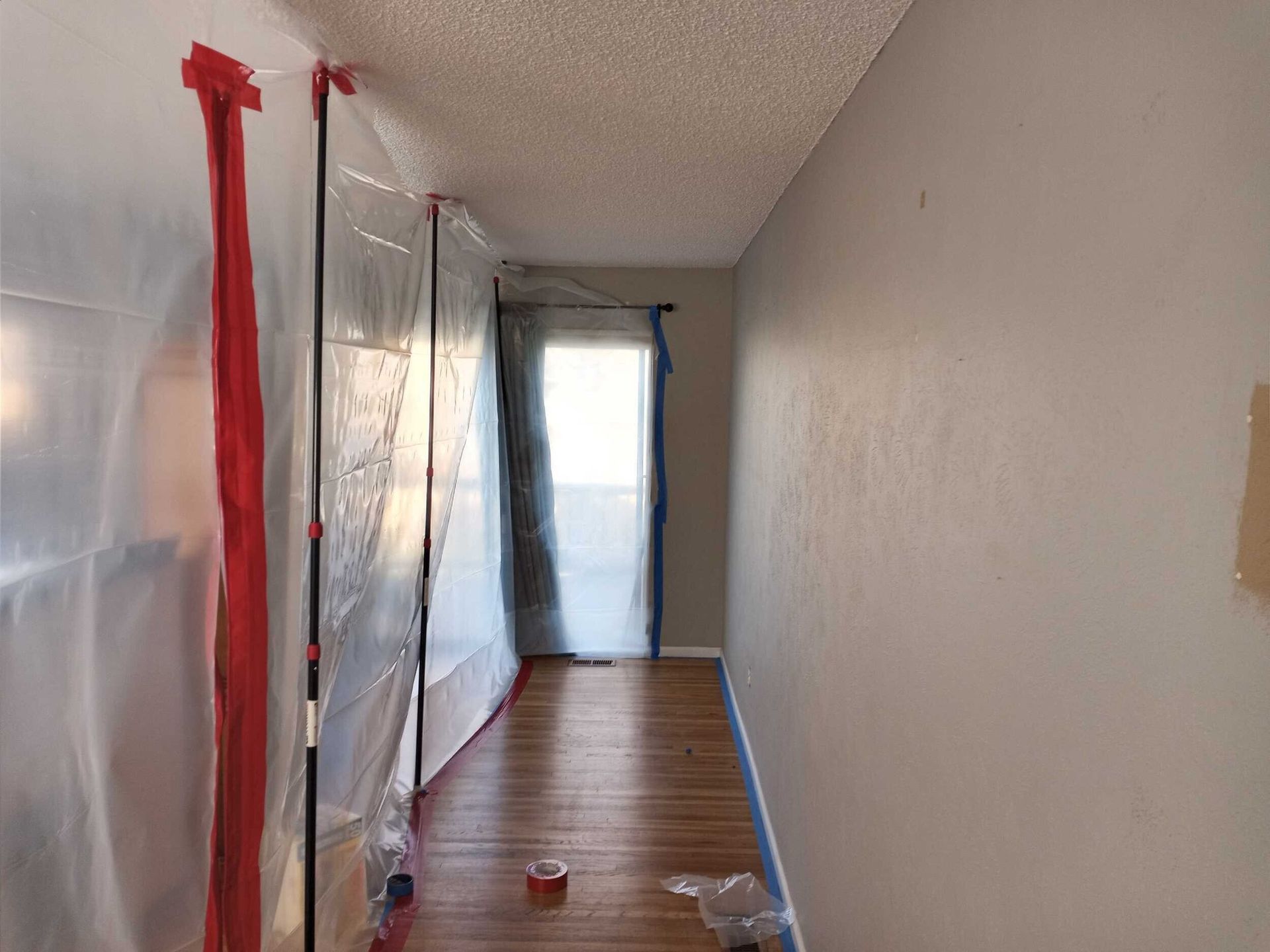 Hallway with plastic sheeting taped to the left wall, wooden floor, door at the end with window.