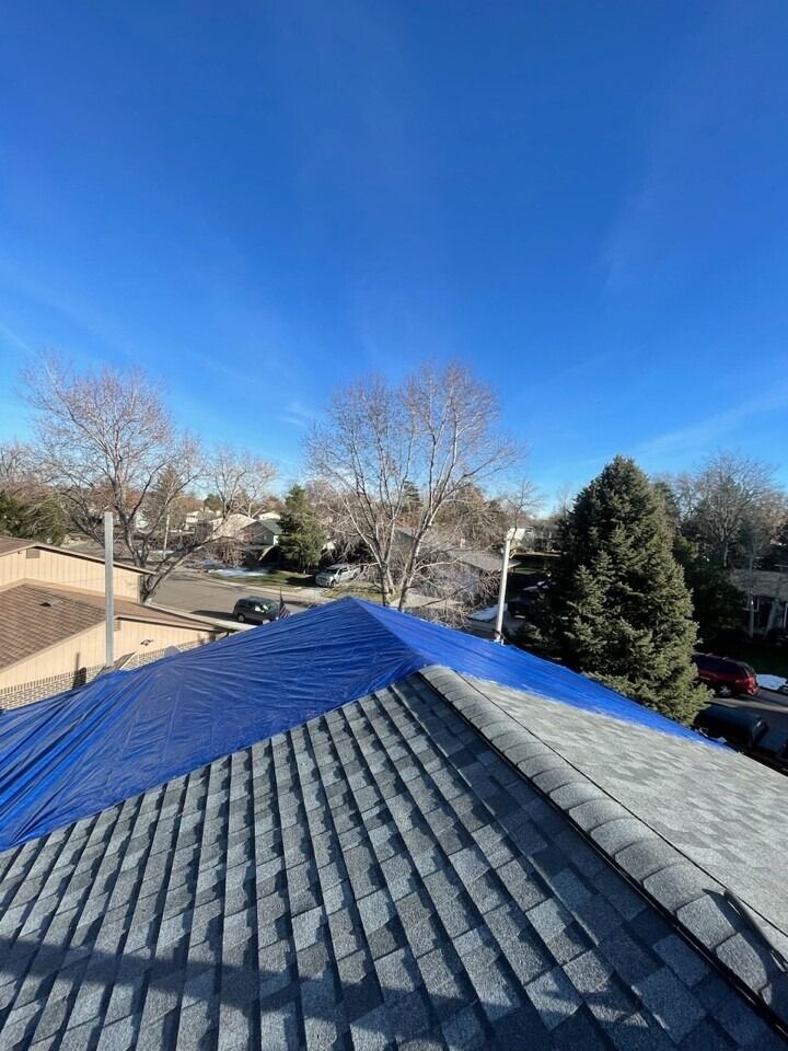 Blue tarp covering a portion of a gray shingled roof under a bright blue sky.