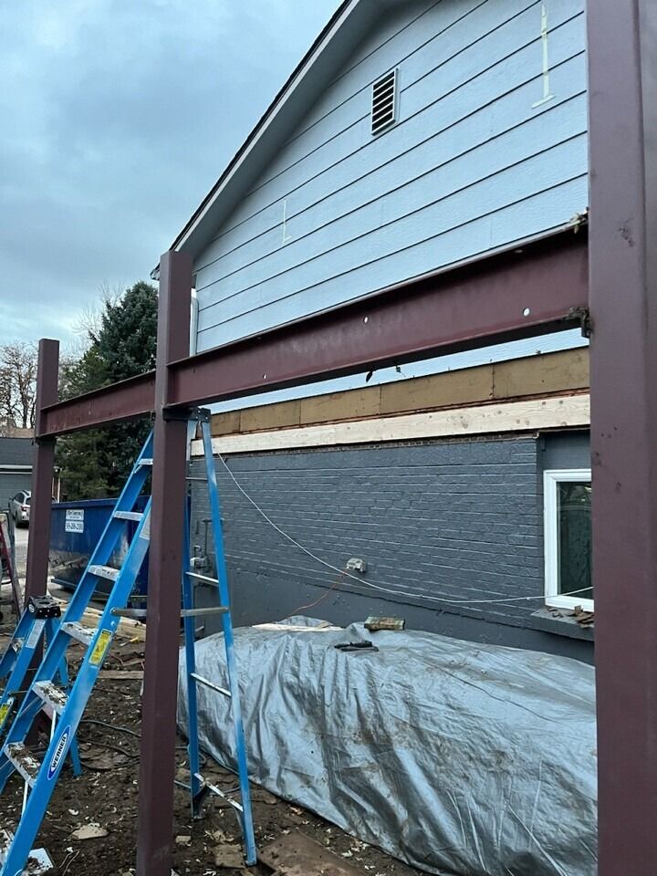 Construction site: steel beams supporting a roof addition on a blue-sided house. Ladders and tarp present.
