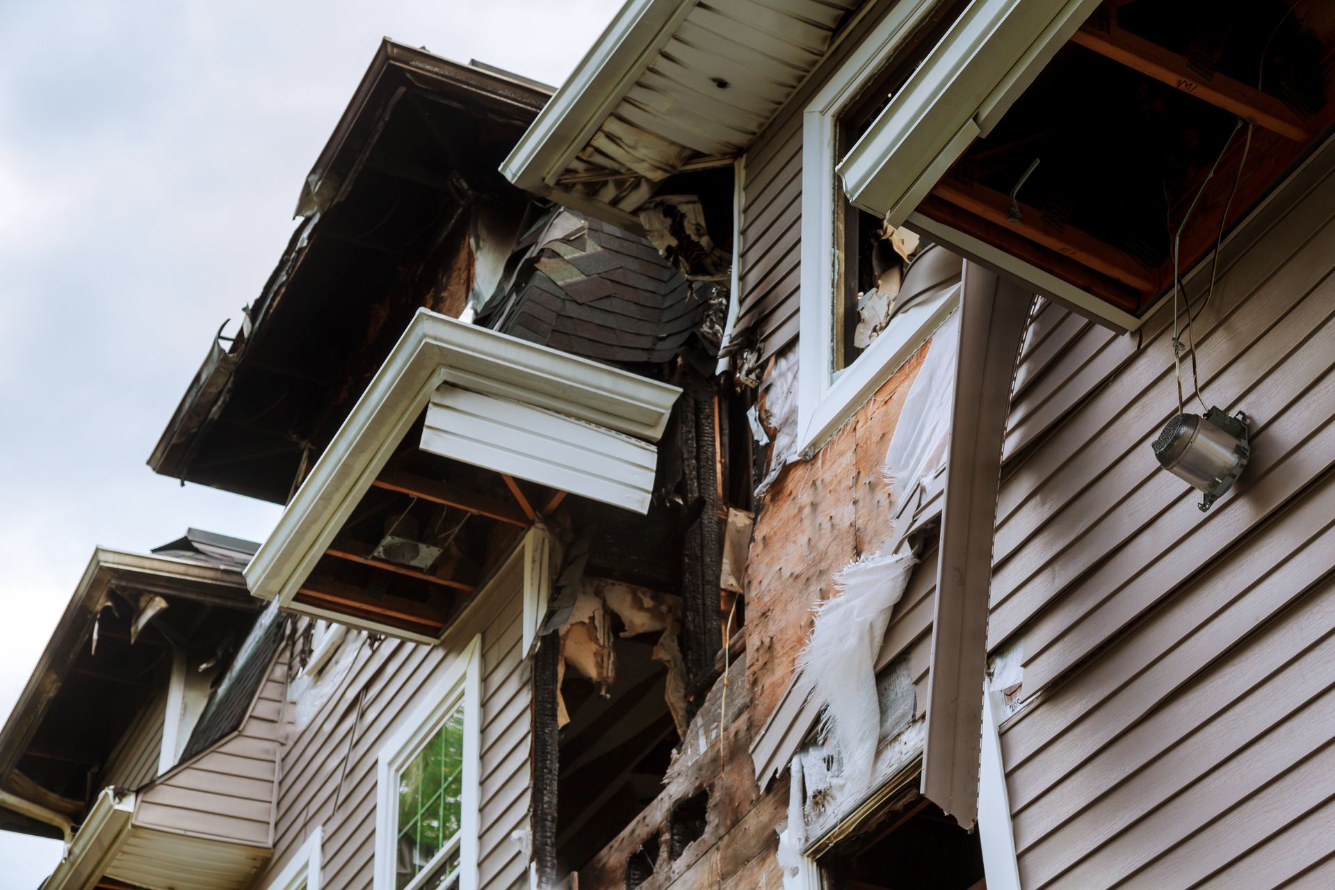 Damaged two-story house exterior after a fire, with visible charring, missing siding, and exposed framing.