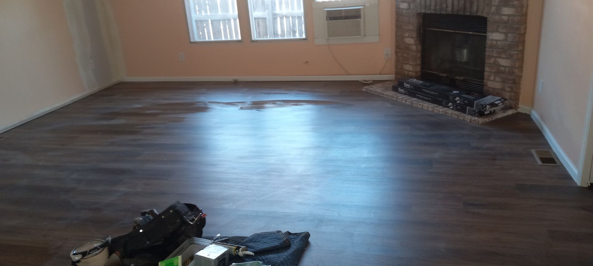 Living room with dark wood-look flooring, fireplace, and windows. Some items sit on the floor.