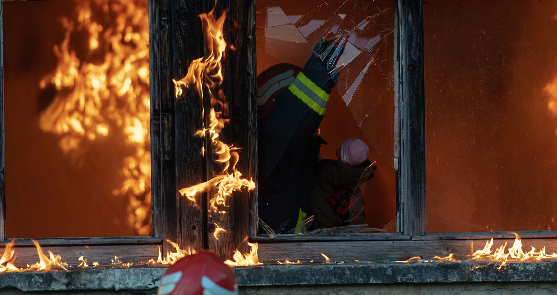 Firefighter breaks through a window of a burning building, flames engulf the structure.
