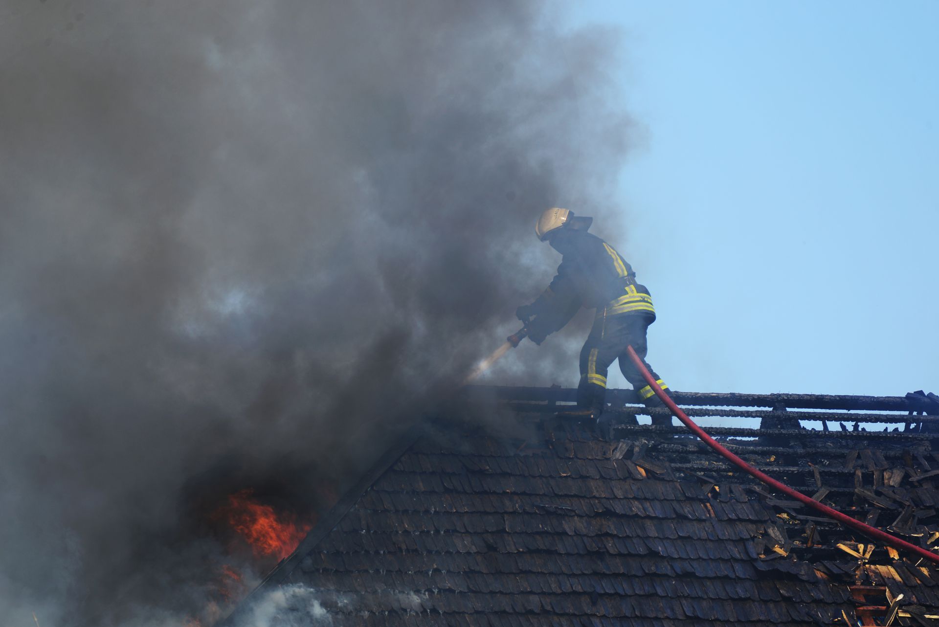 Firefighter on a burning roof, spraying water, with dark smoke billowing.