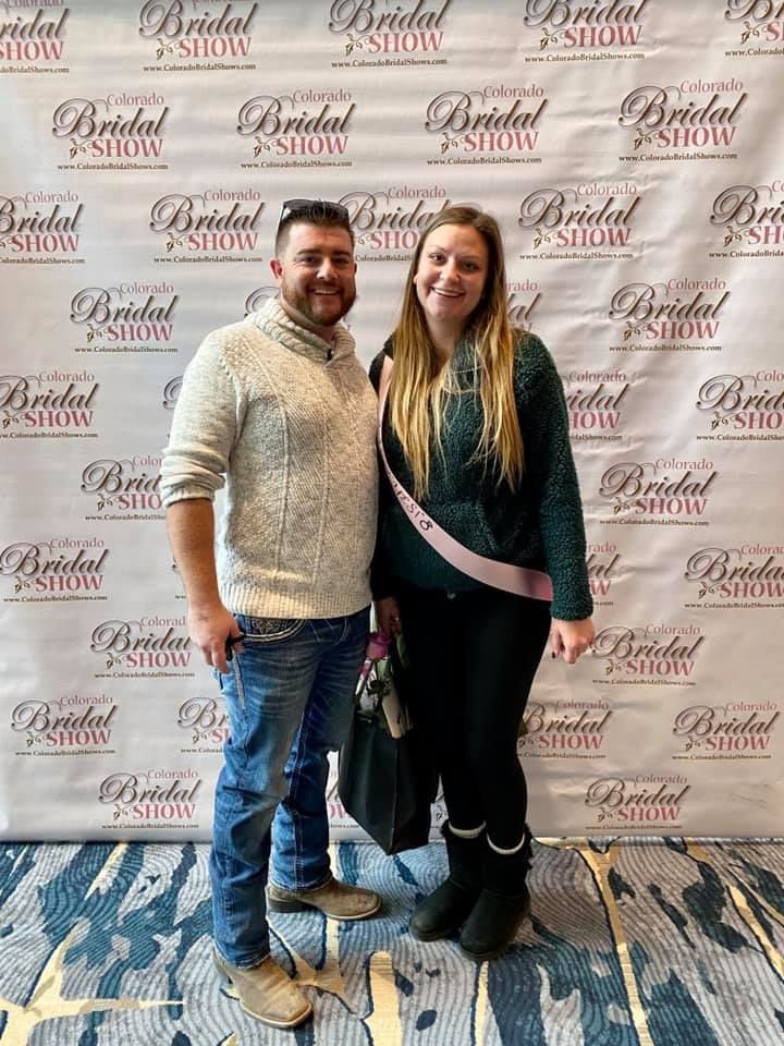 Man and woman at a bridal show, standing in front of a branded backdrop. Woman wears a sash.