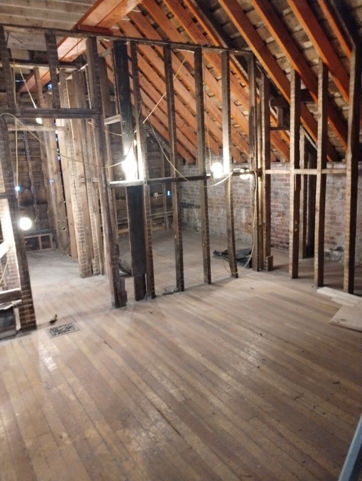 Interior of an attic with exposed wooden beams, studs, brick walls, and hardwood floor.