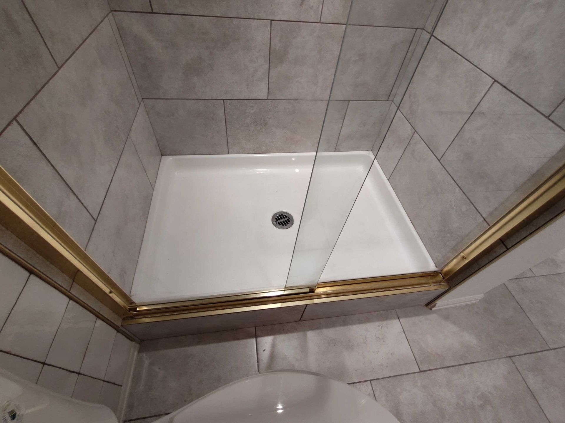 A small shower stall with a white base and a gold-colored frame, surrounded by gray tiled walls.