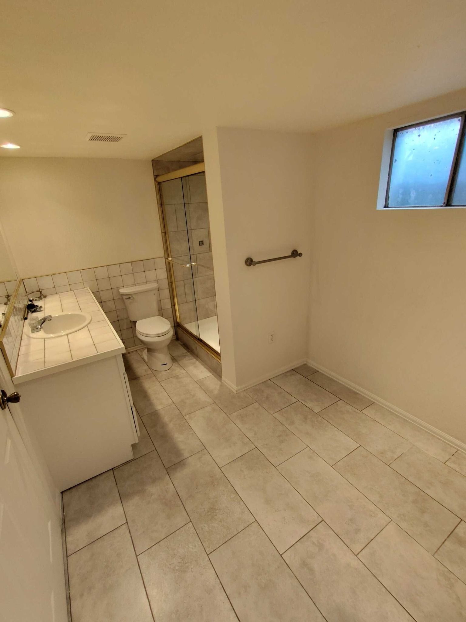 Bathroom with white vanity, toilet, glass shower, and gray tiled floor. Small window on the right.