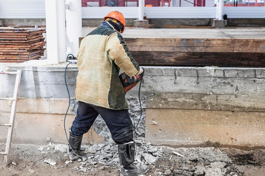 Construction worker in a tan jacket and orange hard hat using a jackhammer on a concrete foundation.