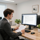 Person reviewing documents at a desk beside a computer in a bright office