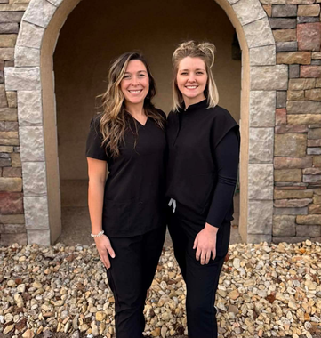 Two women in black scrubs stand under a stone archway.