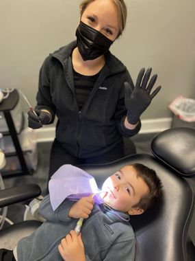 Dental worker in black gloves and mask examining a patient's mouth. The patient has a red beard and glasses.