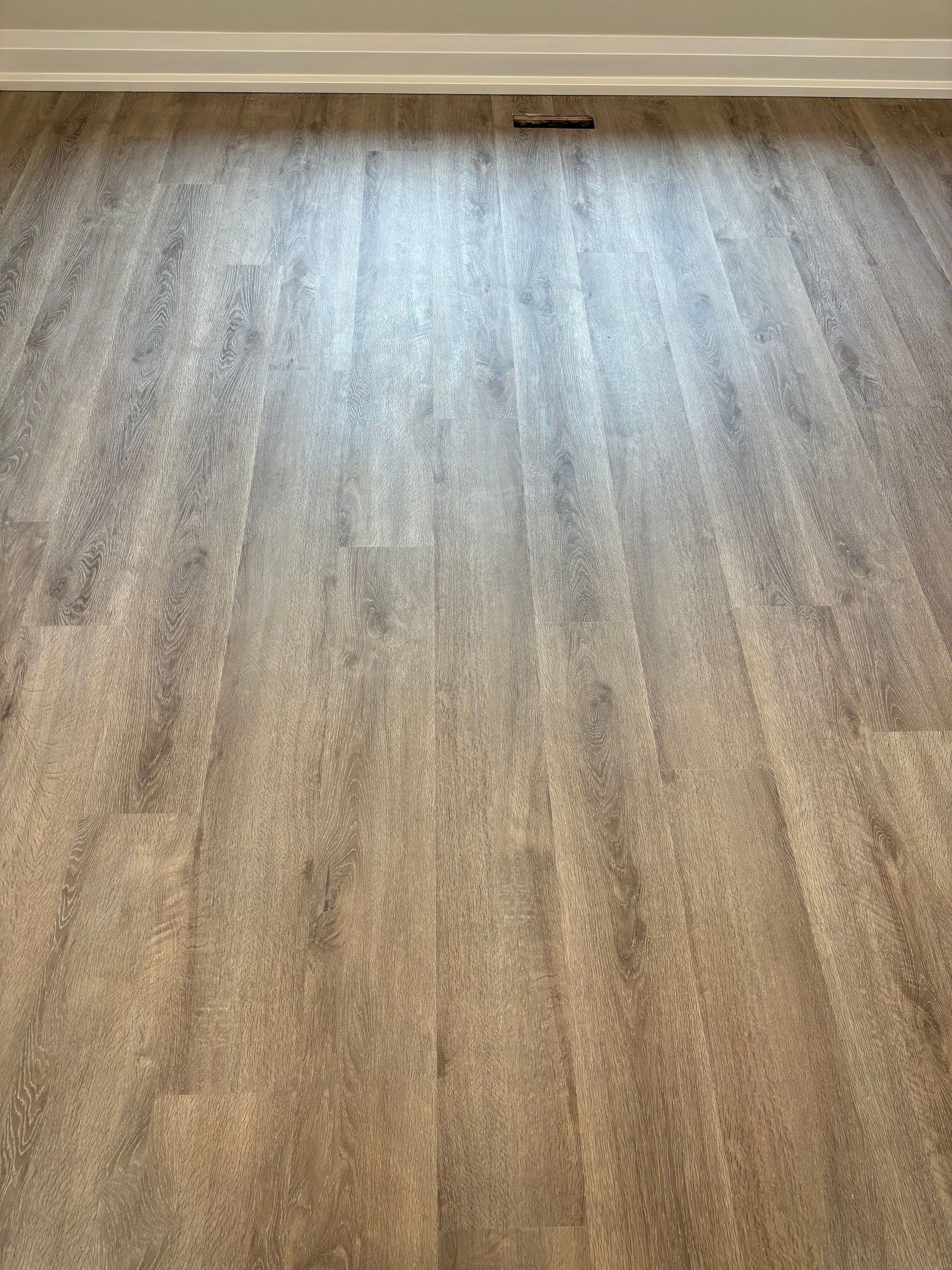 Wood-look flooring in a room. Light brown planks with a textured surface, and a white trim at the top.