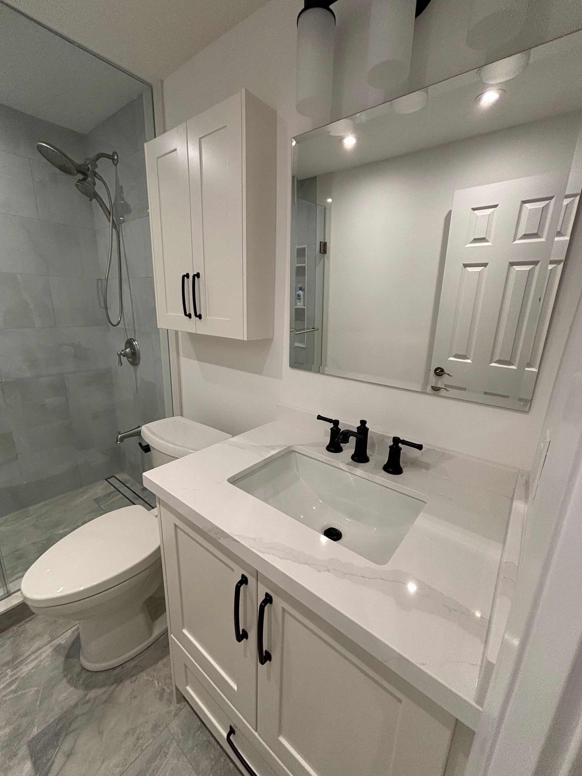 White bathroom with shower, toilet, vanity with black fixtures, and a mirrored cabinet.