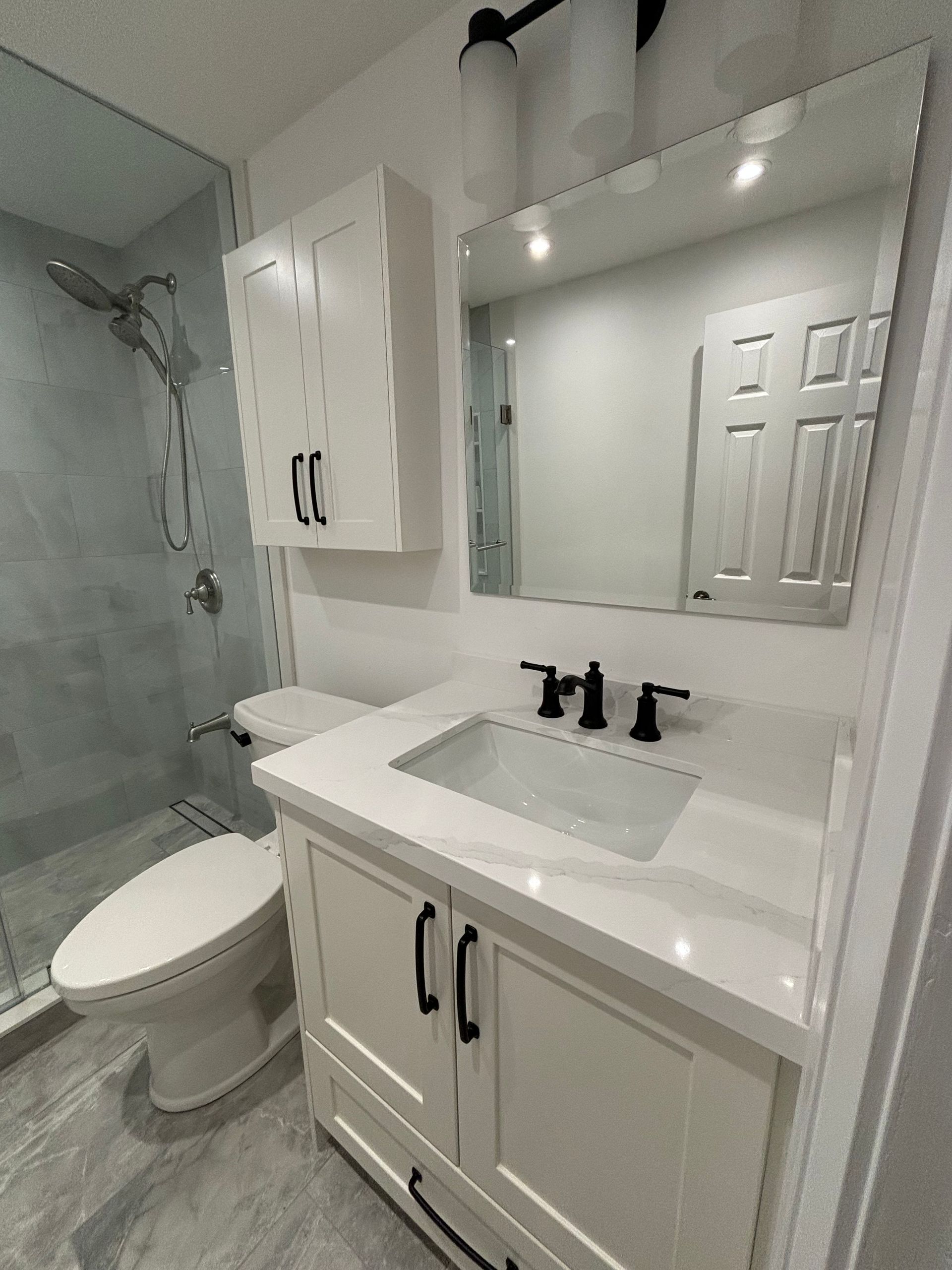 Bathroom with white vanity, sink, mirror, toilet, shower with glass door. Gray tile.