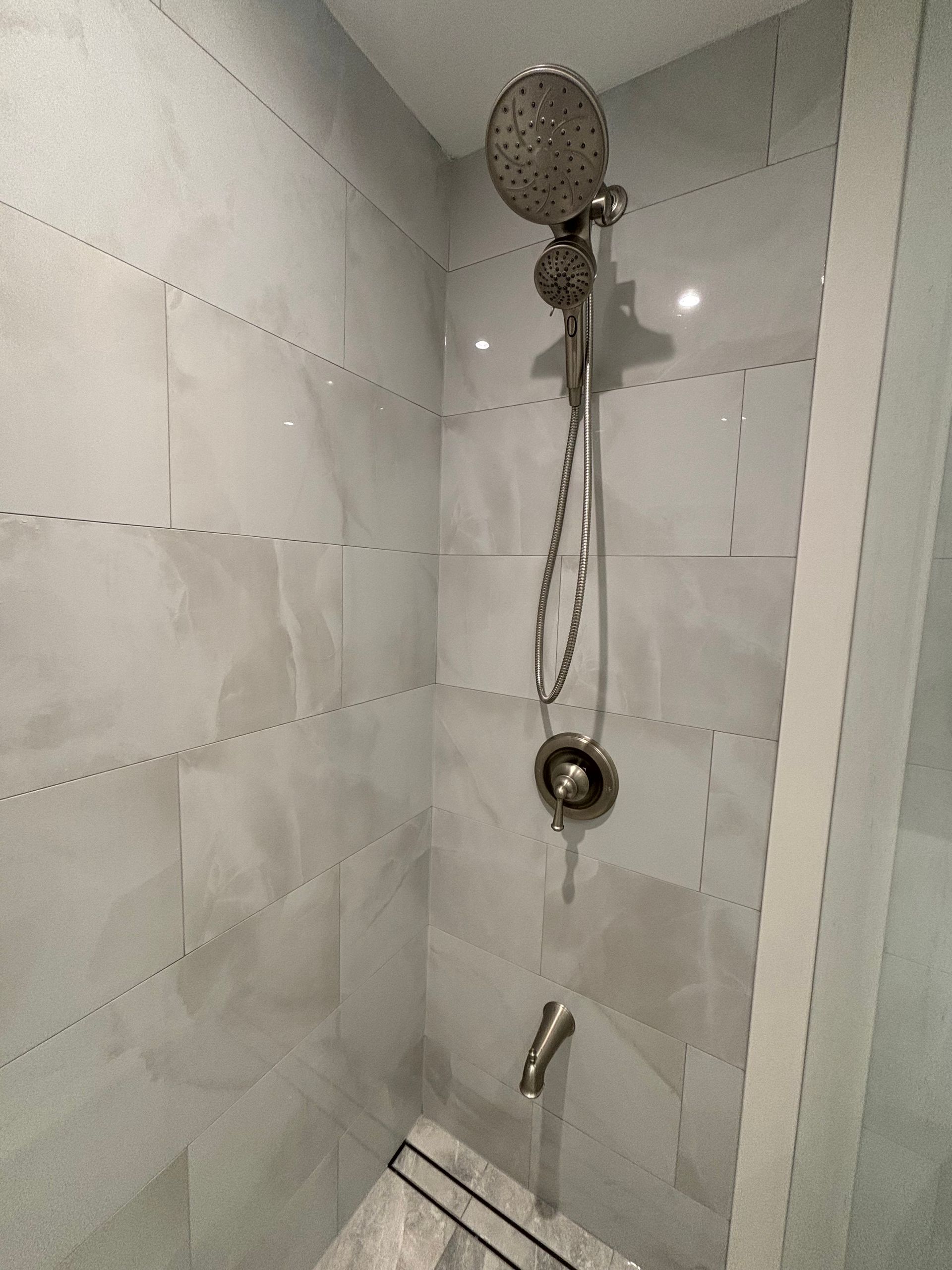 Shower with gray and white tile, showerhead, handheld sprayer, and faucet.