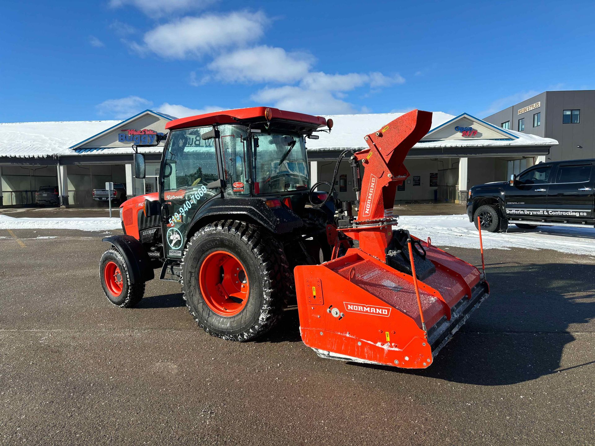 Red tractor with front snow blower parked in a snowy lot outside buildings