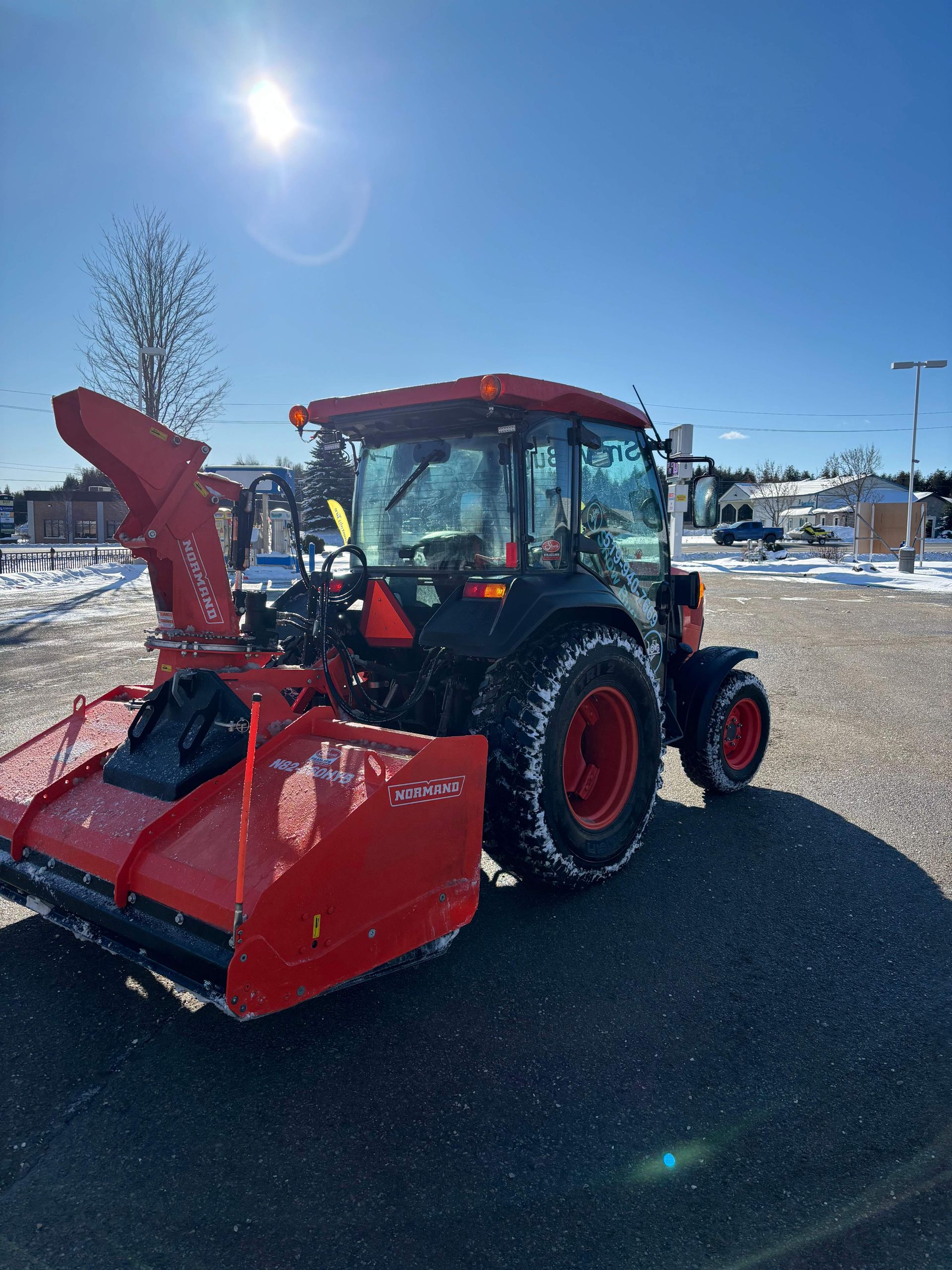 Red tractor with front loader and snowblower attachment parked on a snowy lot under a bright blue sky.