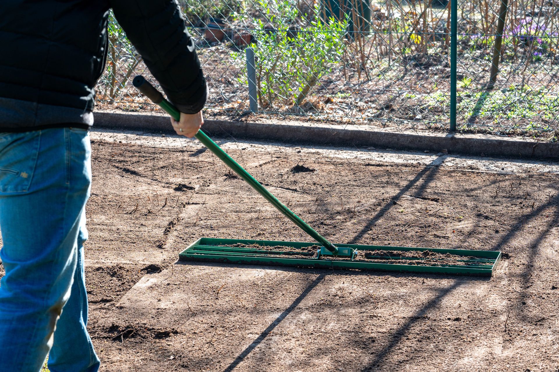 Person raking dirt with a green rake in a fenced outdoor area