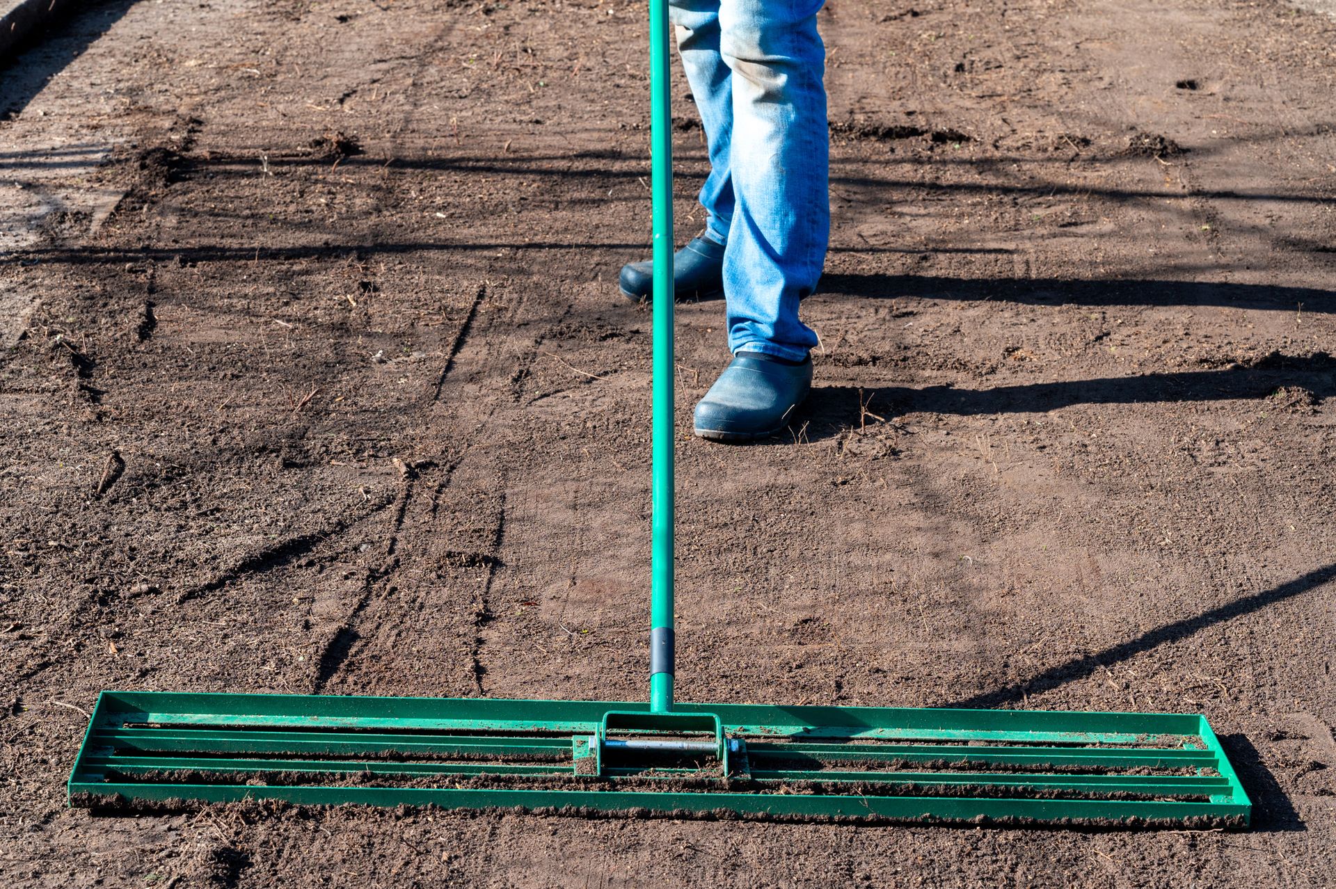 Person using a green rake to clear a cracked dirt surface outdoors