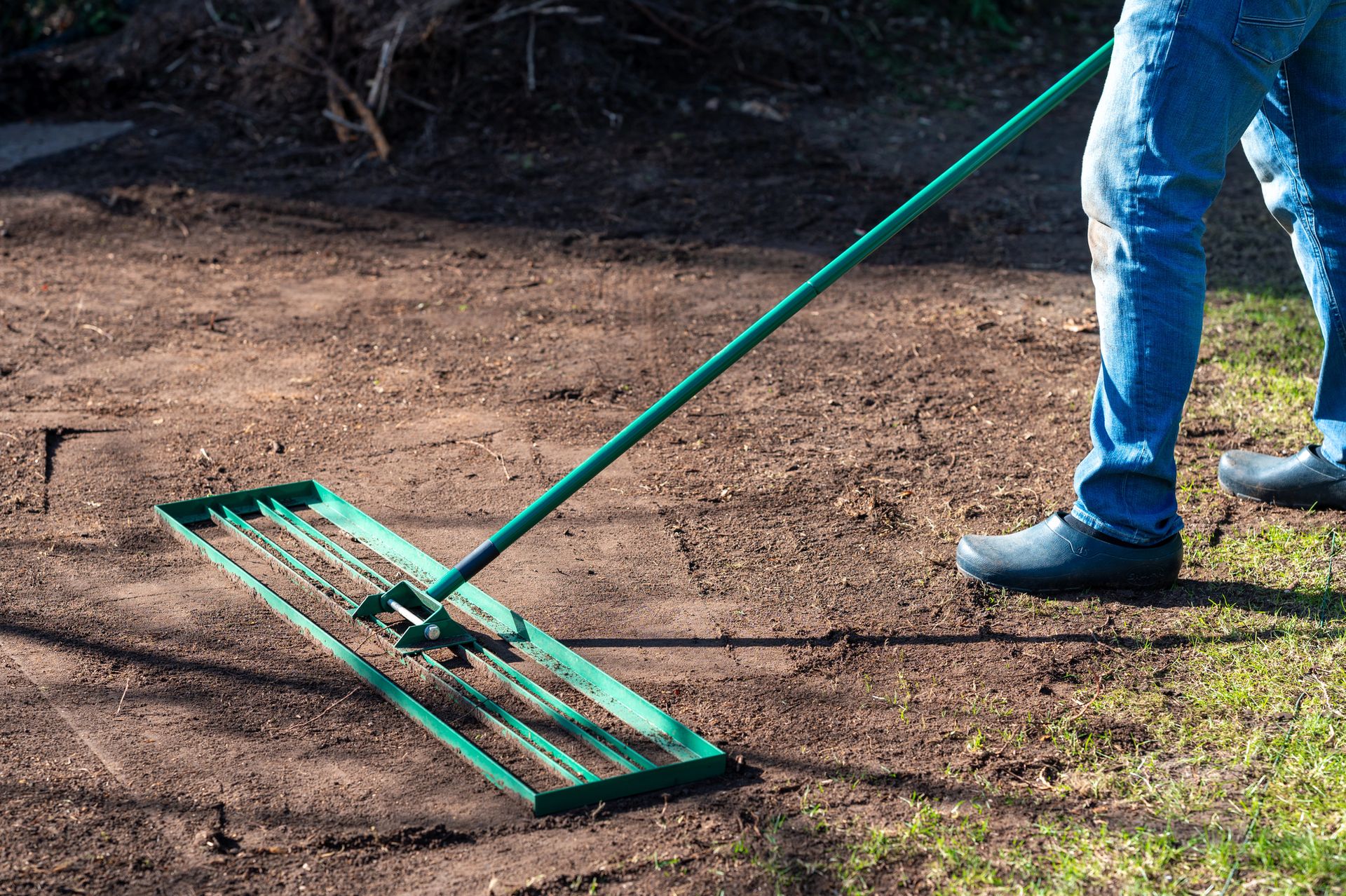 Person using a green rake to level dirt on a garden path beside grass in blue jeans and boots