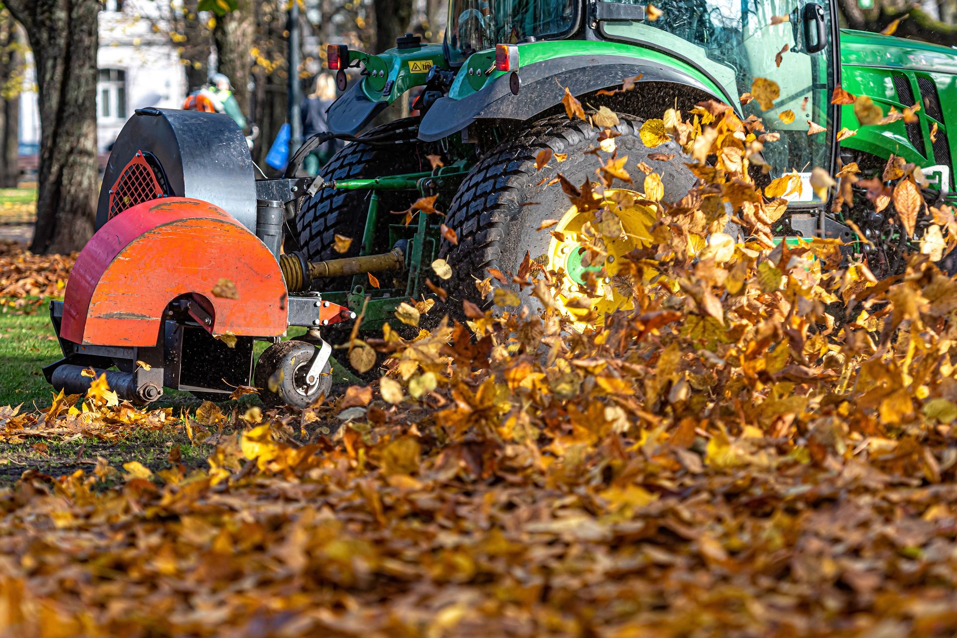 Leaf blower clearing autumn leaves on a street with an orange machine and green tractor