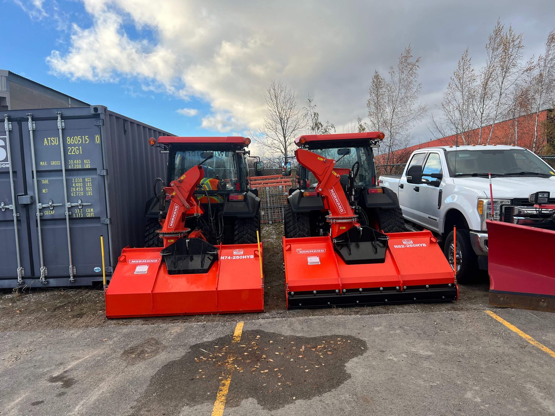 Two red snowplow attachments parked beside shipping containers and a white van in a lot.