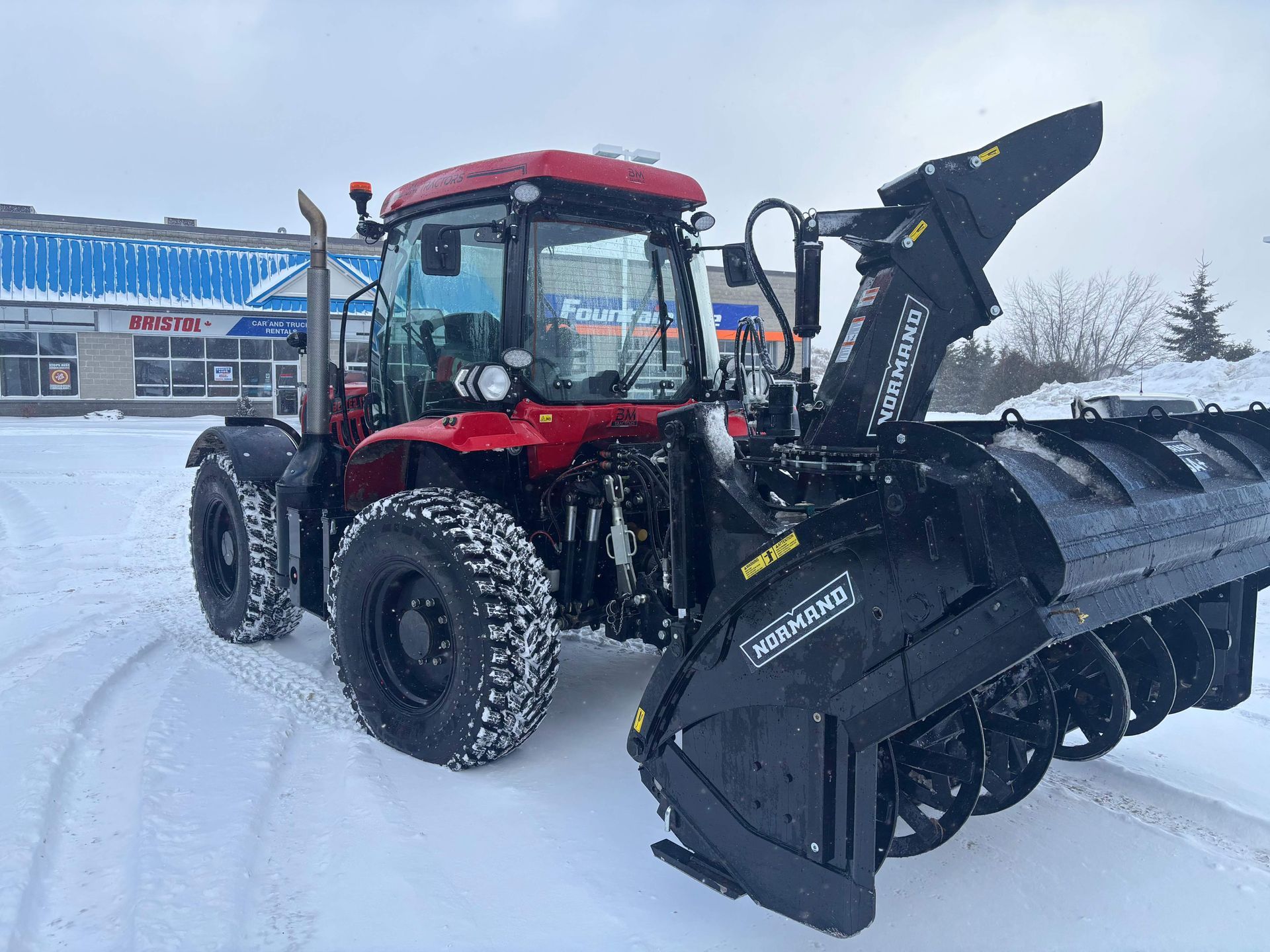 Red tractor with snow plow attachment on a snowy road near a blue building