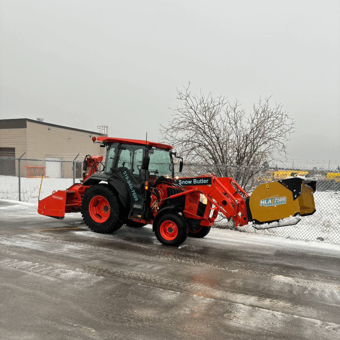 Red tractor with yellow snow blower clearing a snowy driveway beside a building