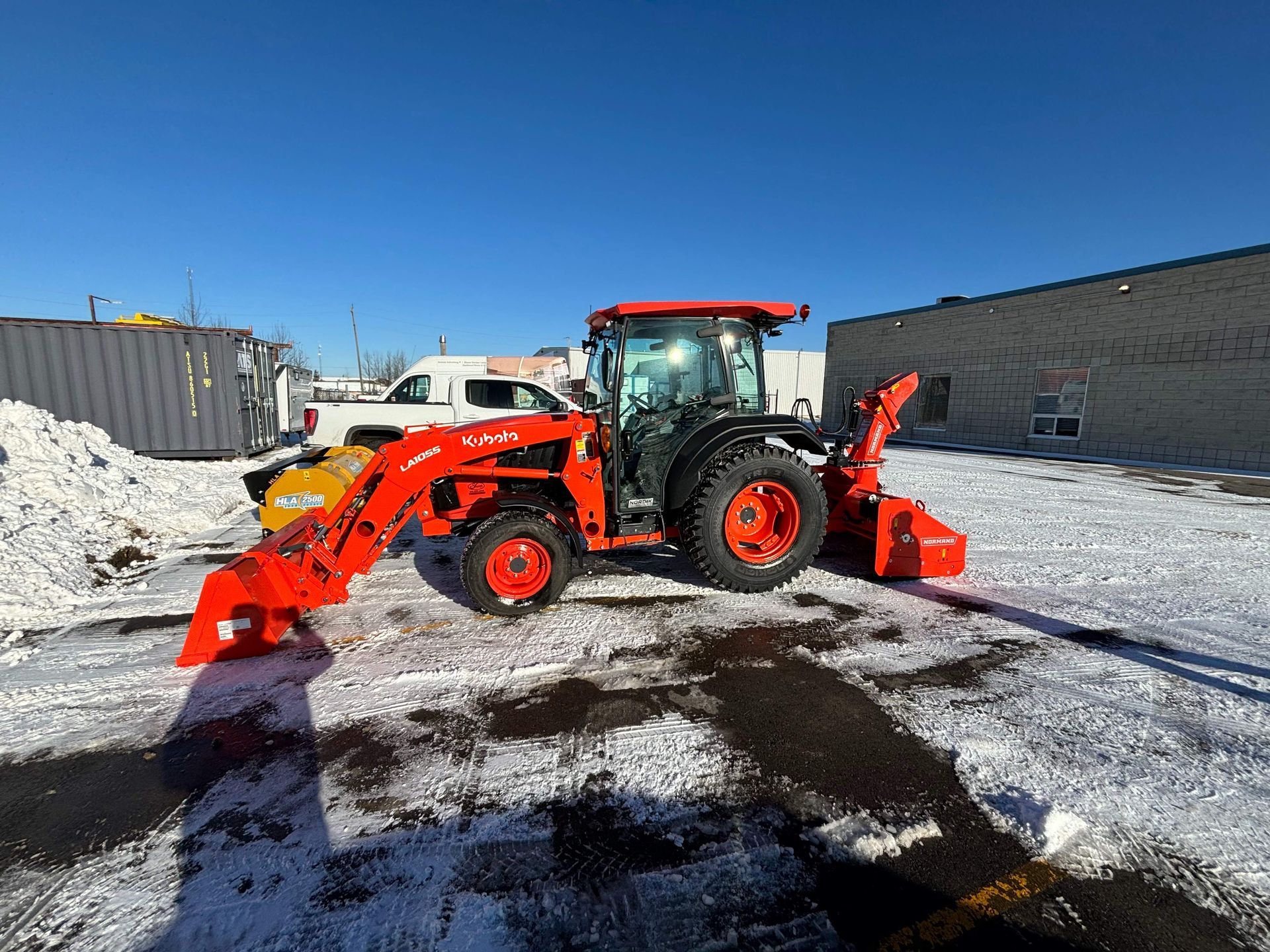 Orange tractor with front loader and rear attachment parked on a snowy lot under a clear blue sky