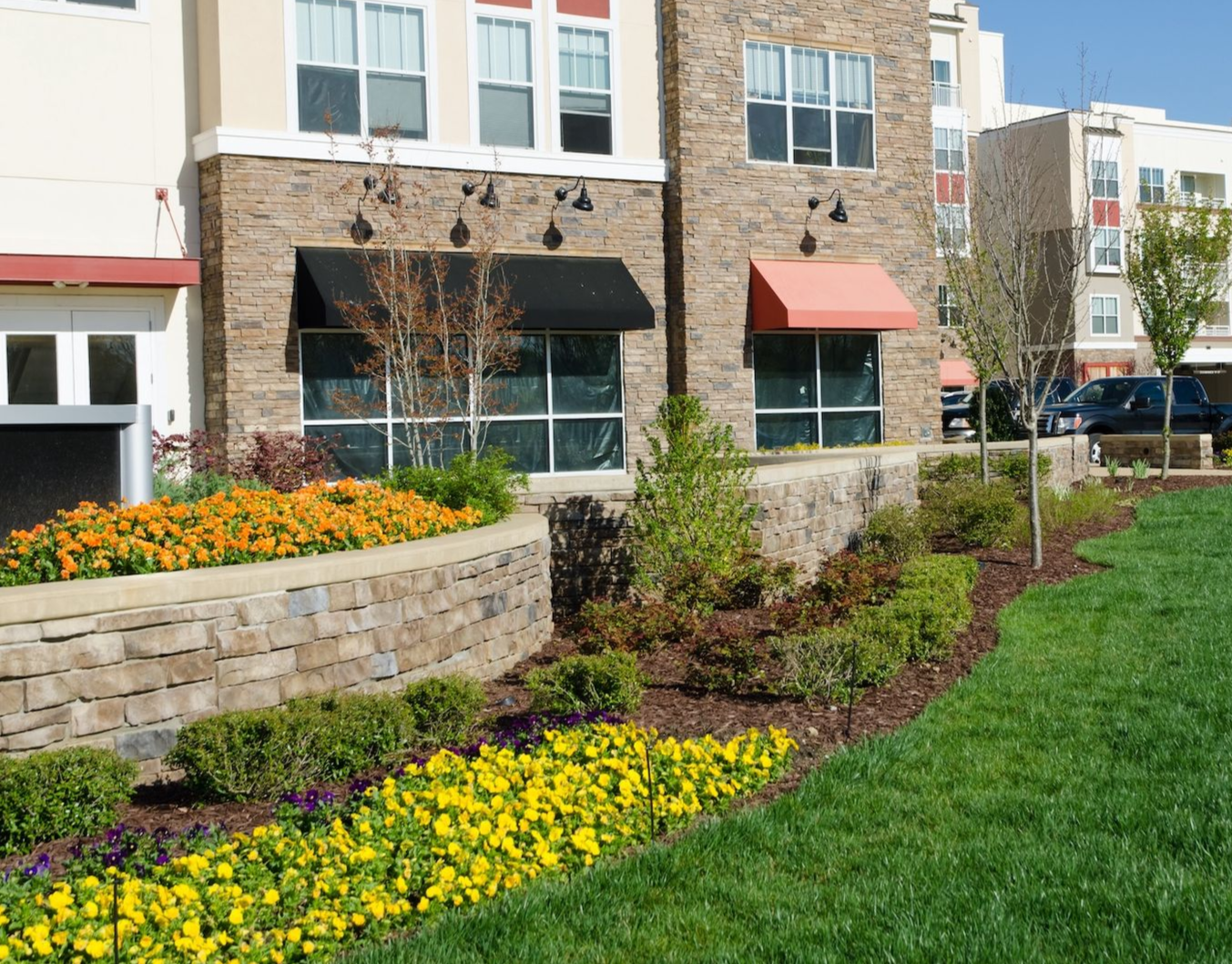 Landscaped apartment courtyard with flower beds, stone planter, and grass in front of brick buildings