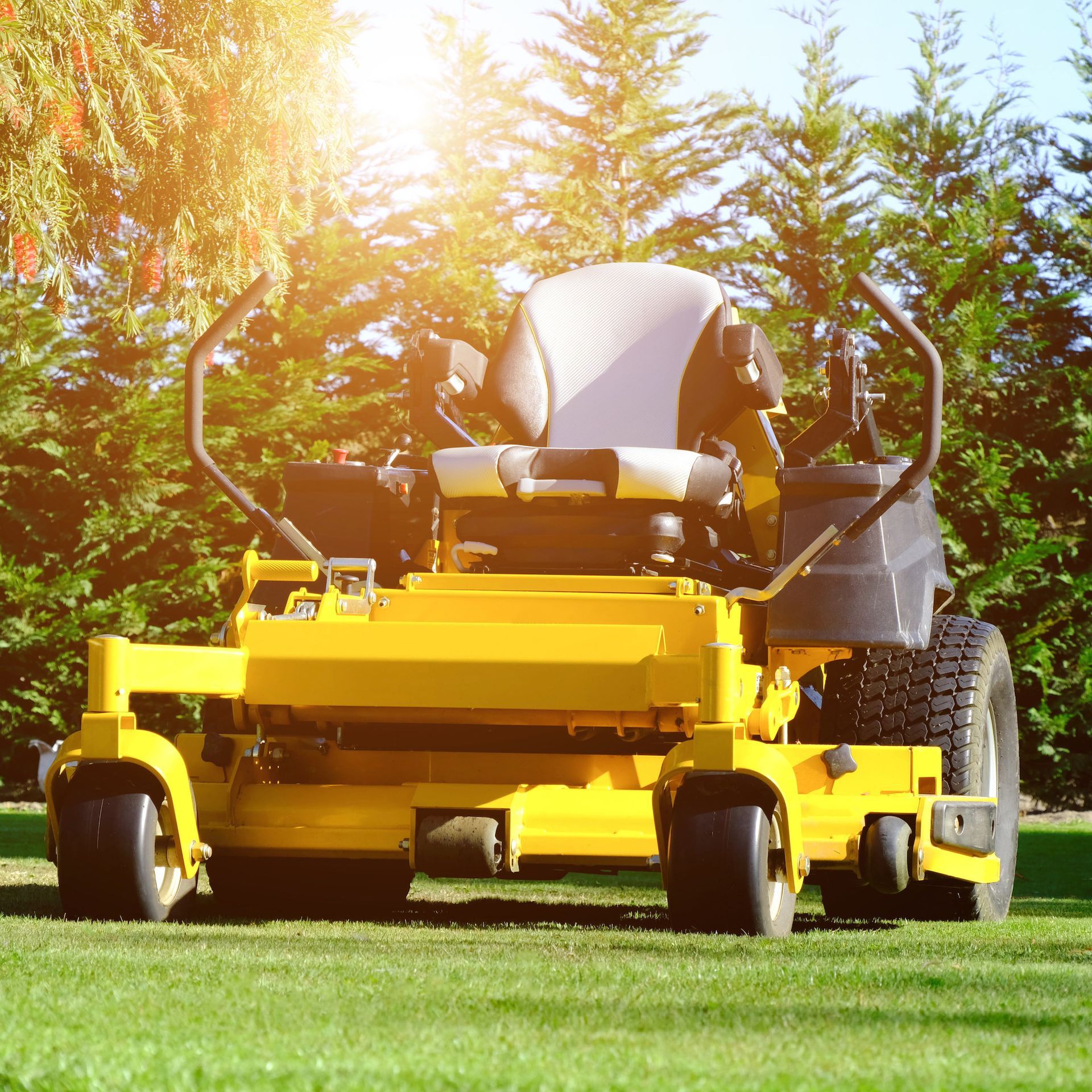 Yellow riding lawn mower on green grass with trees and sunlight in the background
