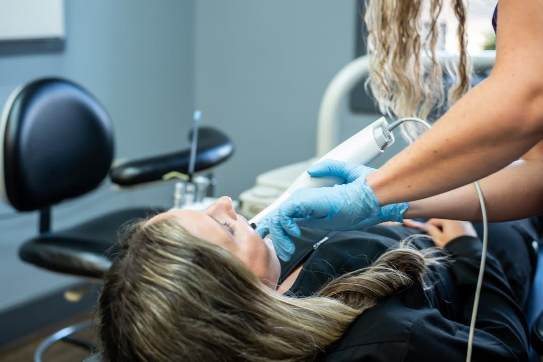 Woman in dentist chair receiving treatment; gloved hand holding tool, light-colored hair, gray walls.
