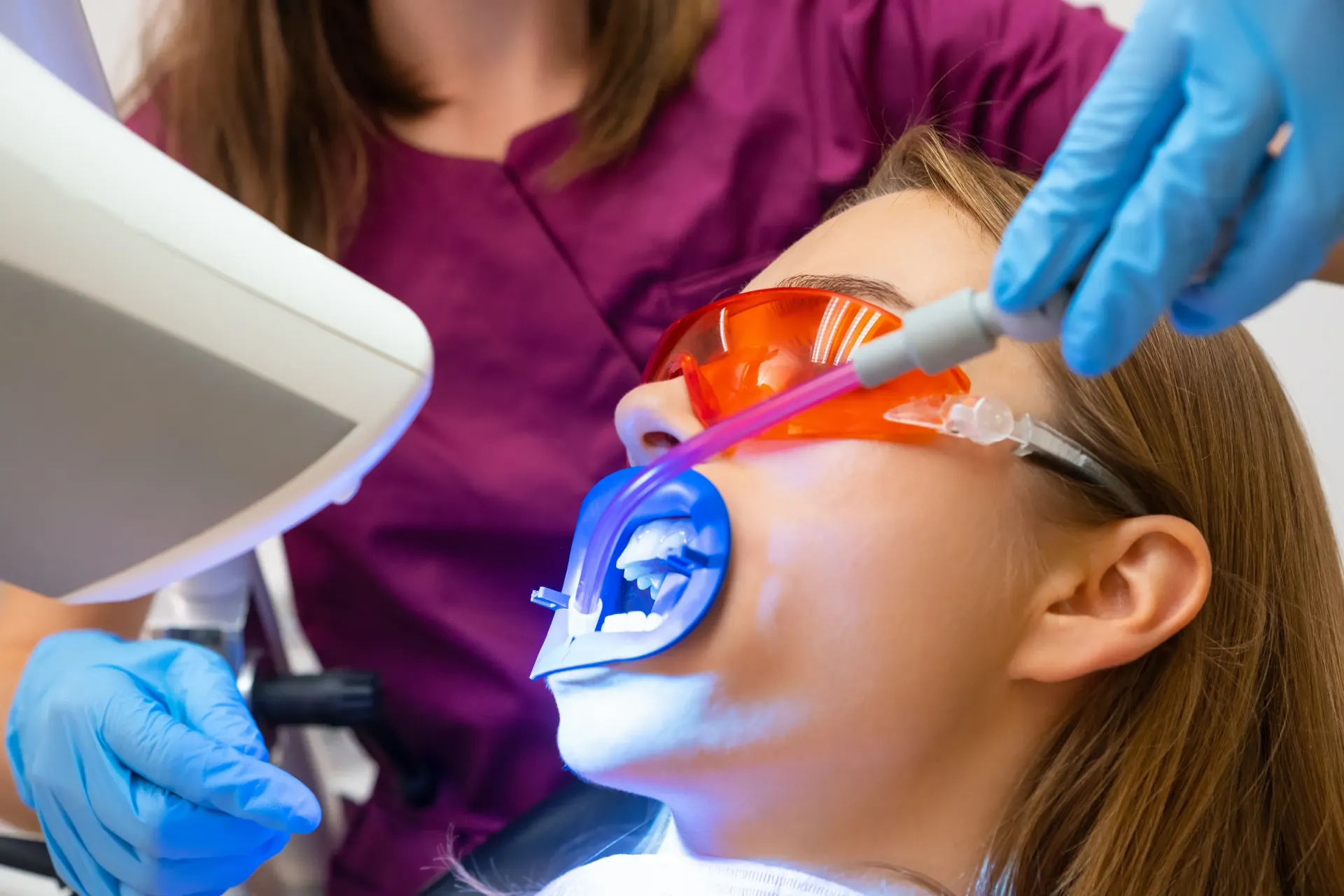 Dentist using a laser on a patient's teeth. Patient wearing orange glasses, blue gloves, purple shirt.