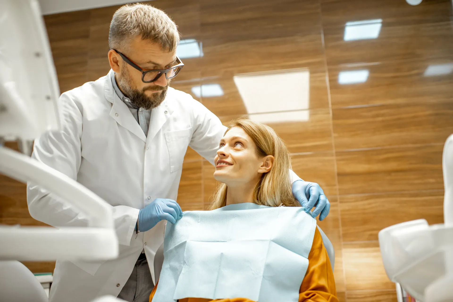 Dentist in lab coat puts bib on patient in dental chair.