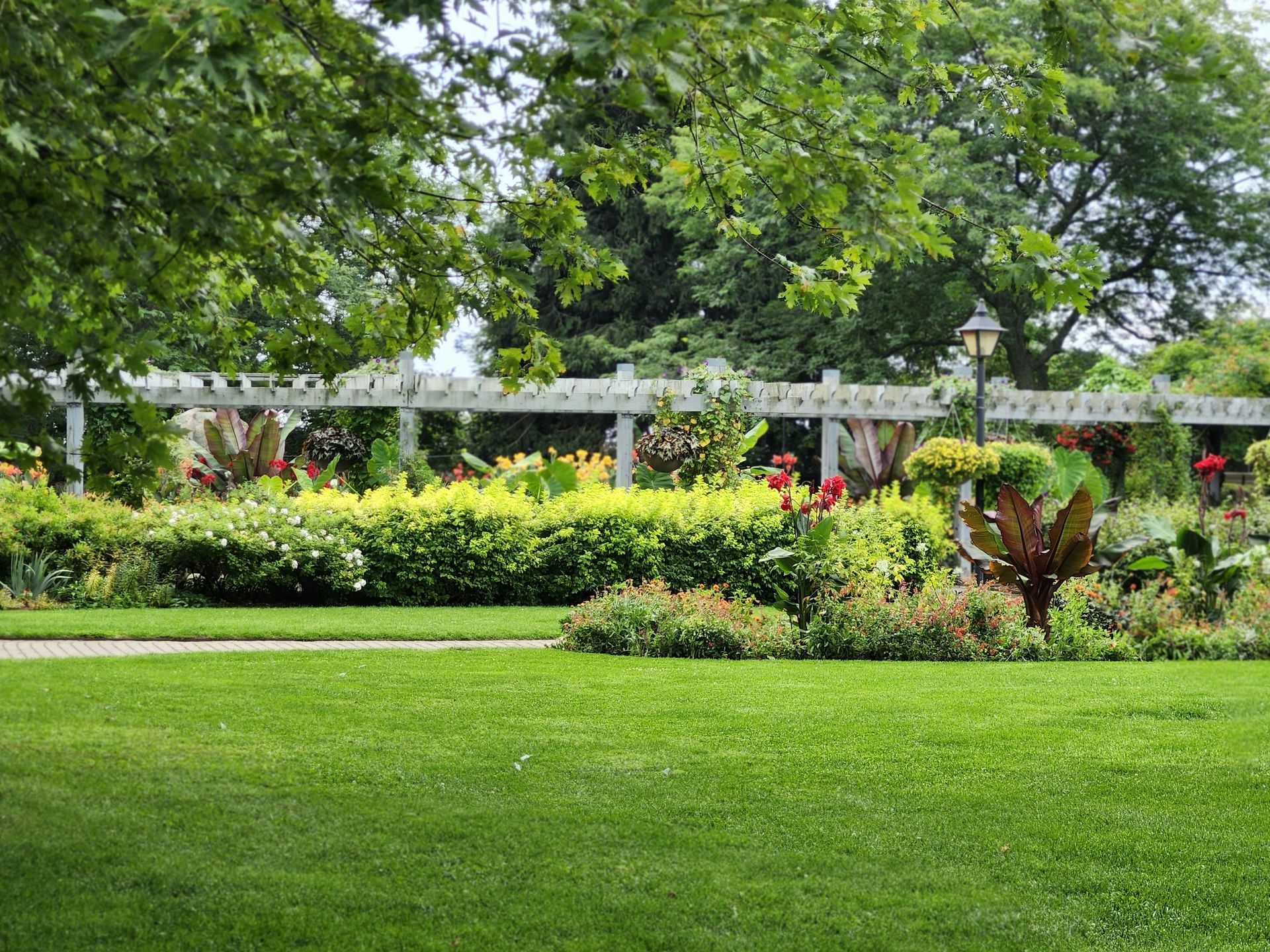 Lush green garden with manicured lawn, hedges, flowering shrubs, and a white pergola under trees