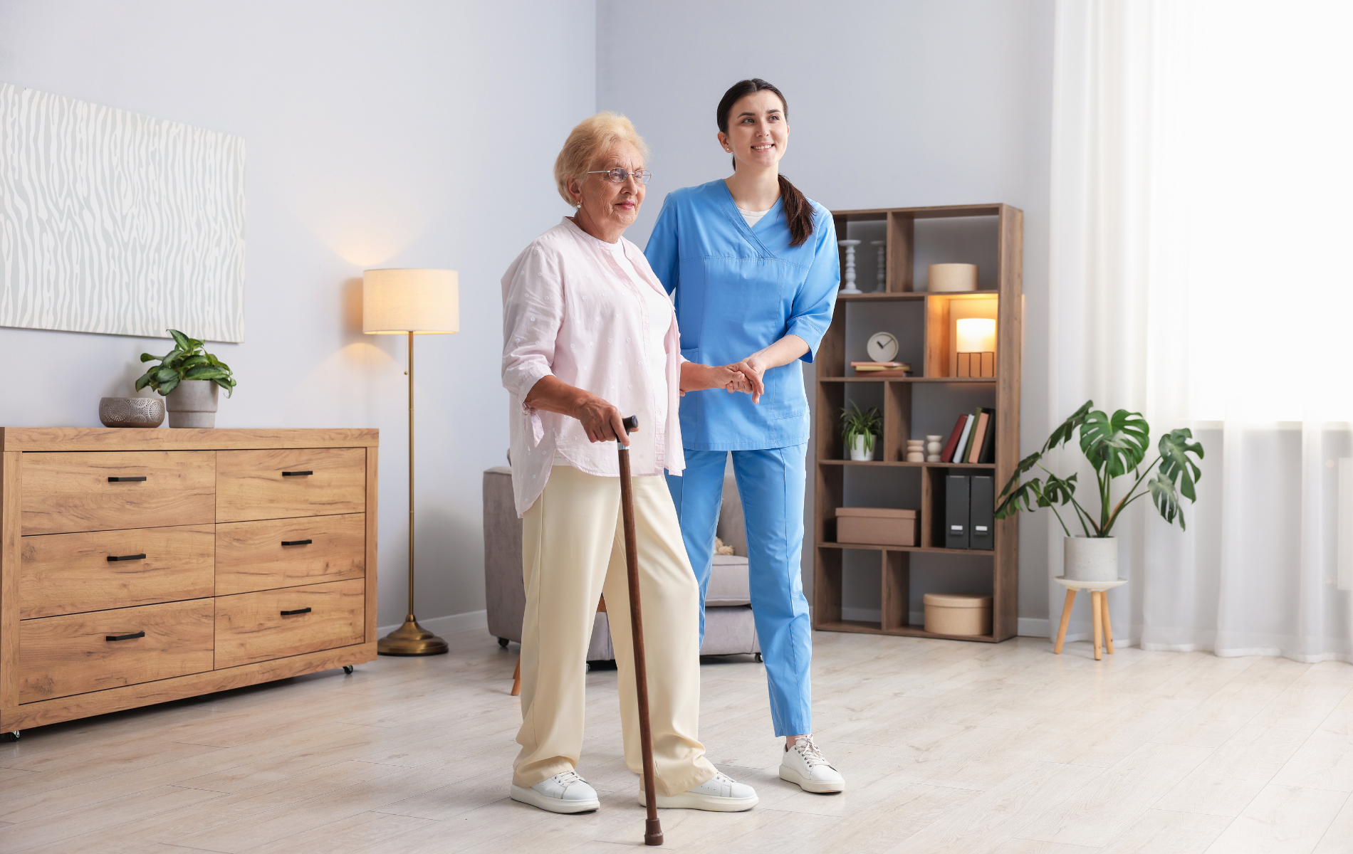 An older adult uses a cane with assistance from a caregiver in a well-lit living room.
