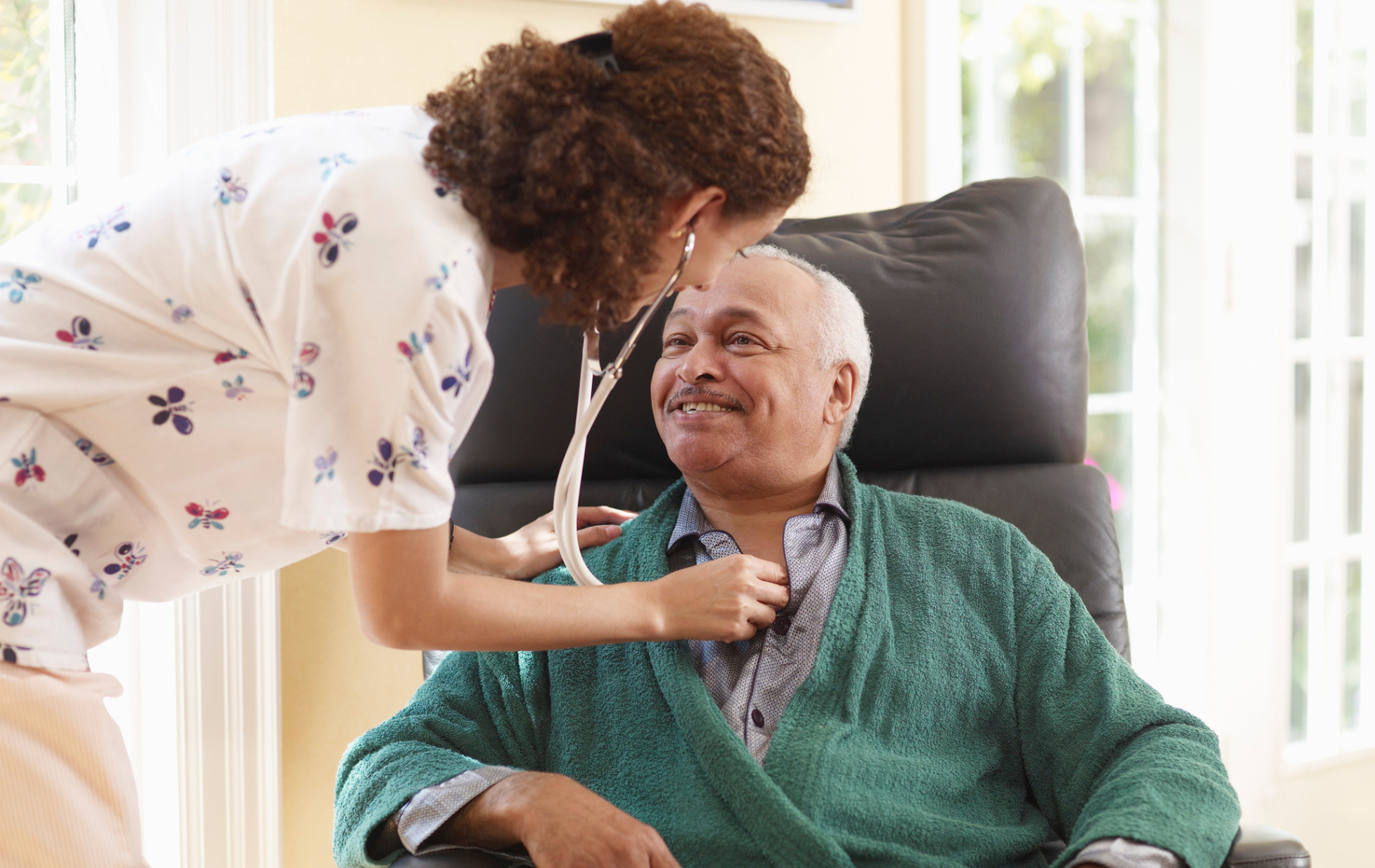 Nurse listens to patient's heart with stethoscope; patient smiles, seated in a chair, wearing a green sweater.
